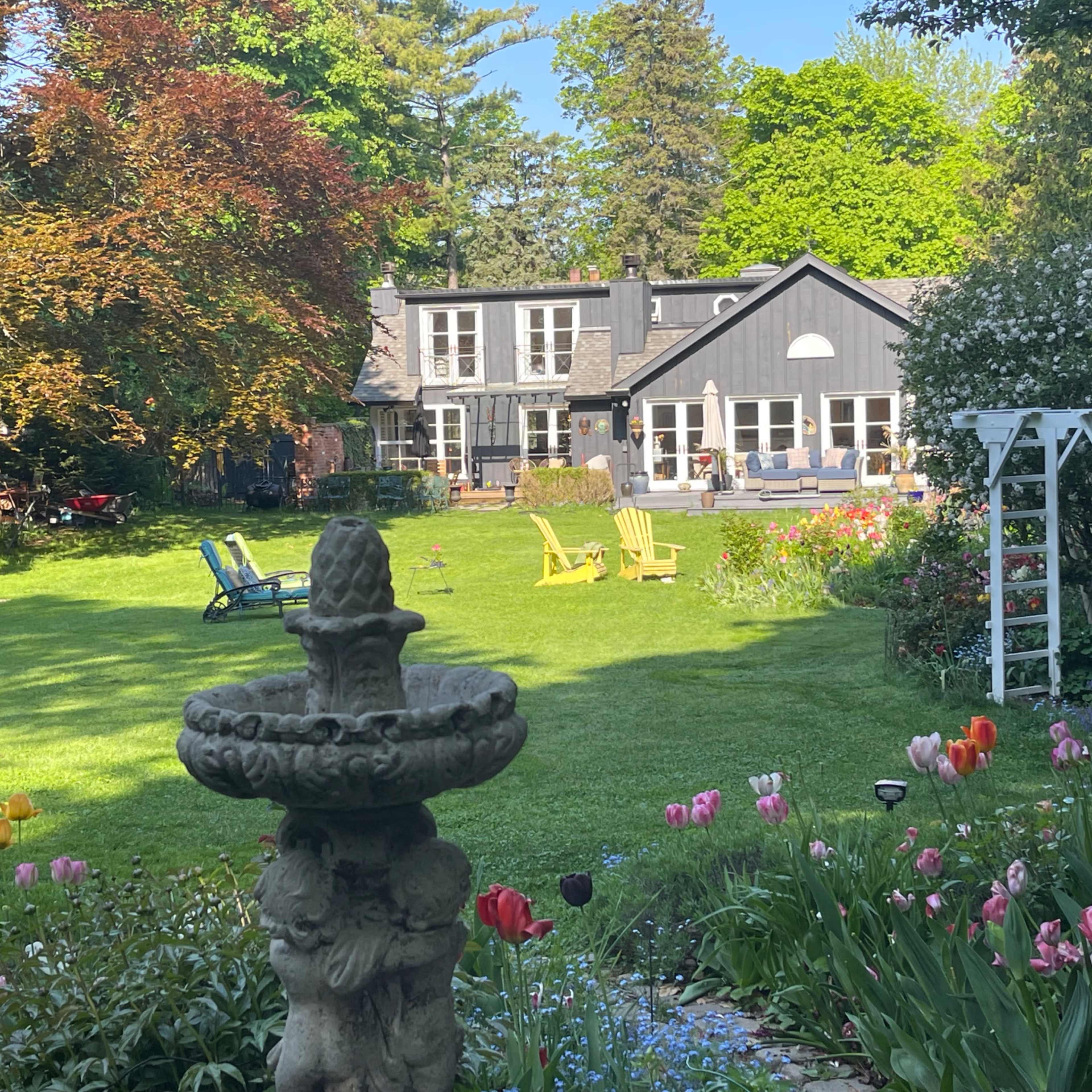 A well-maintained garden features colorful flowers and a stone fountain, with a house and seating area visible in the background.