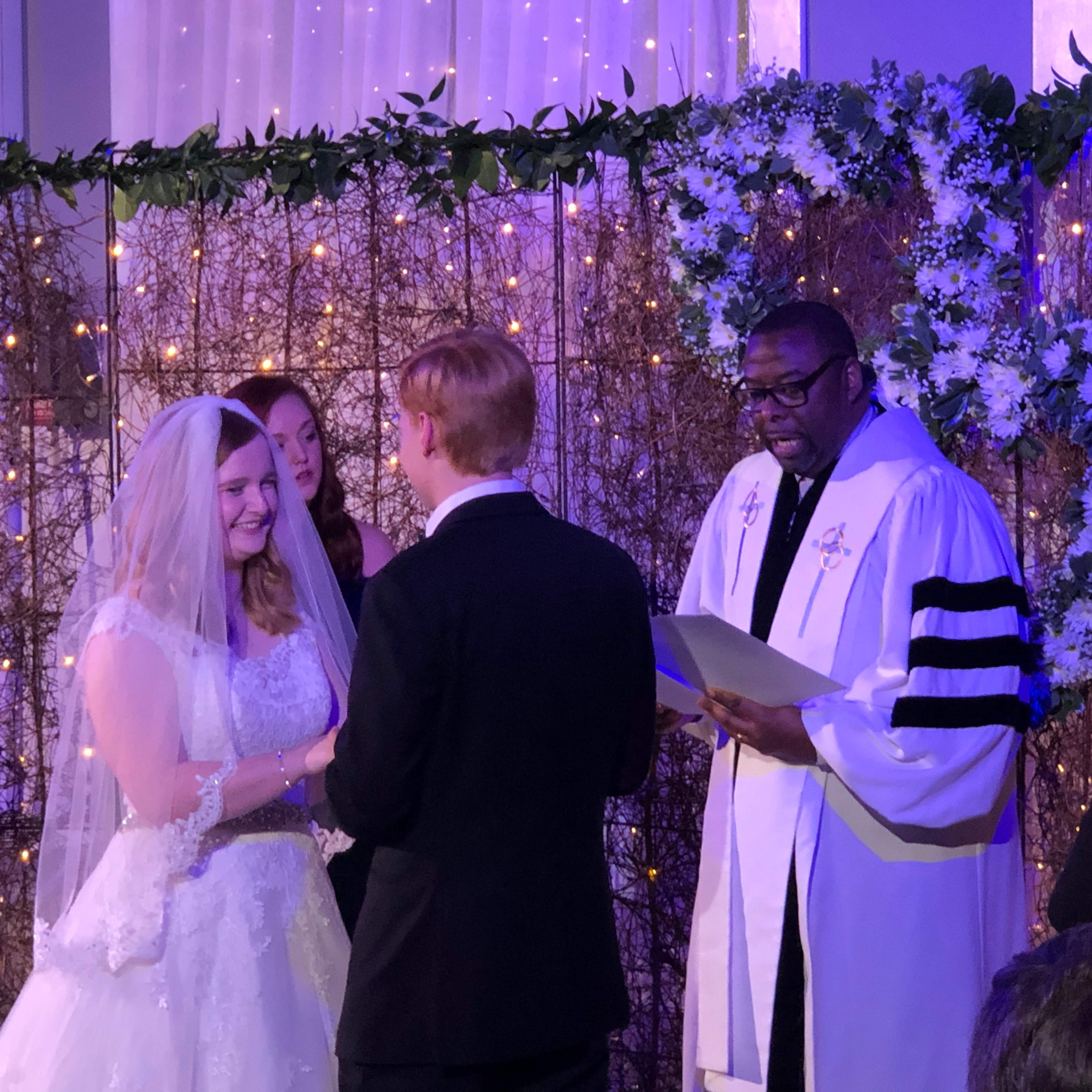A couple exchanges vows during their wedding ceremony, surrounded by attendants and a officiant.