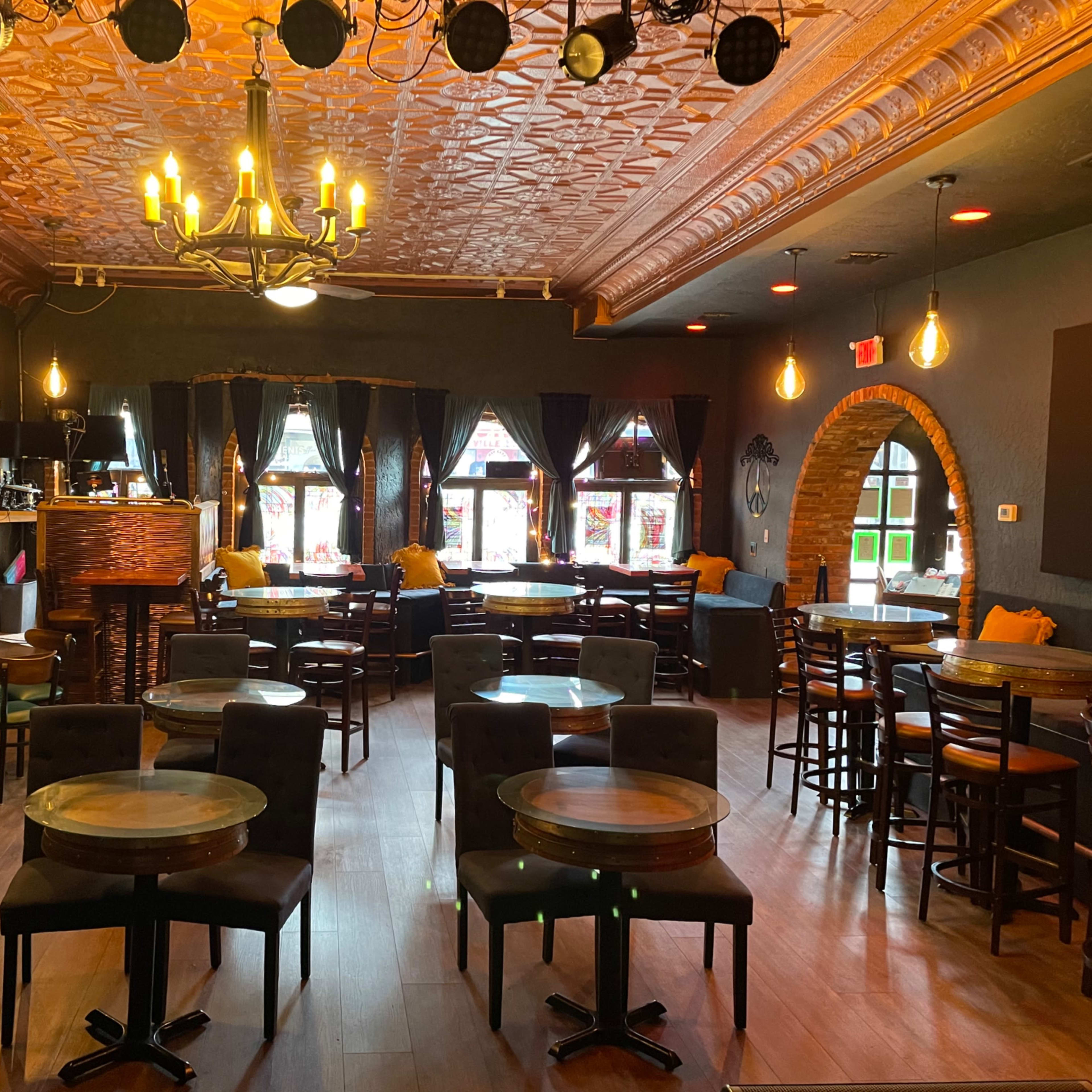 A dimly lit bar area with wooden tables and chairs, adorned with chandeliers and vintage decor, featuring large windows.