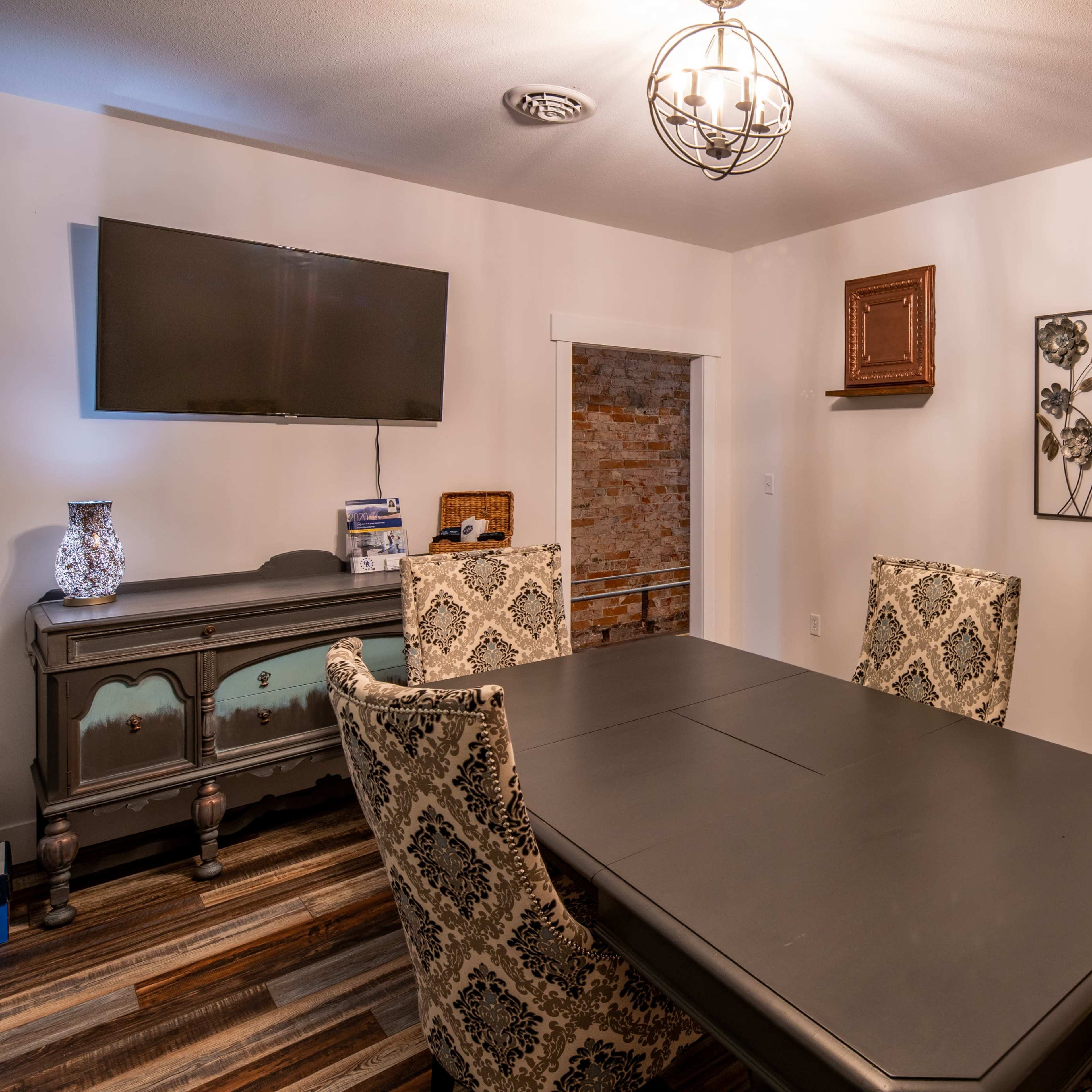 A dining room with a large black table surrounded by ornate chairs, a television mounted on the wall, and decorative elements including framed art and a cabinet.