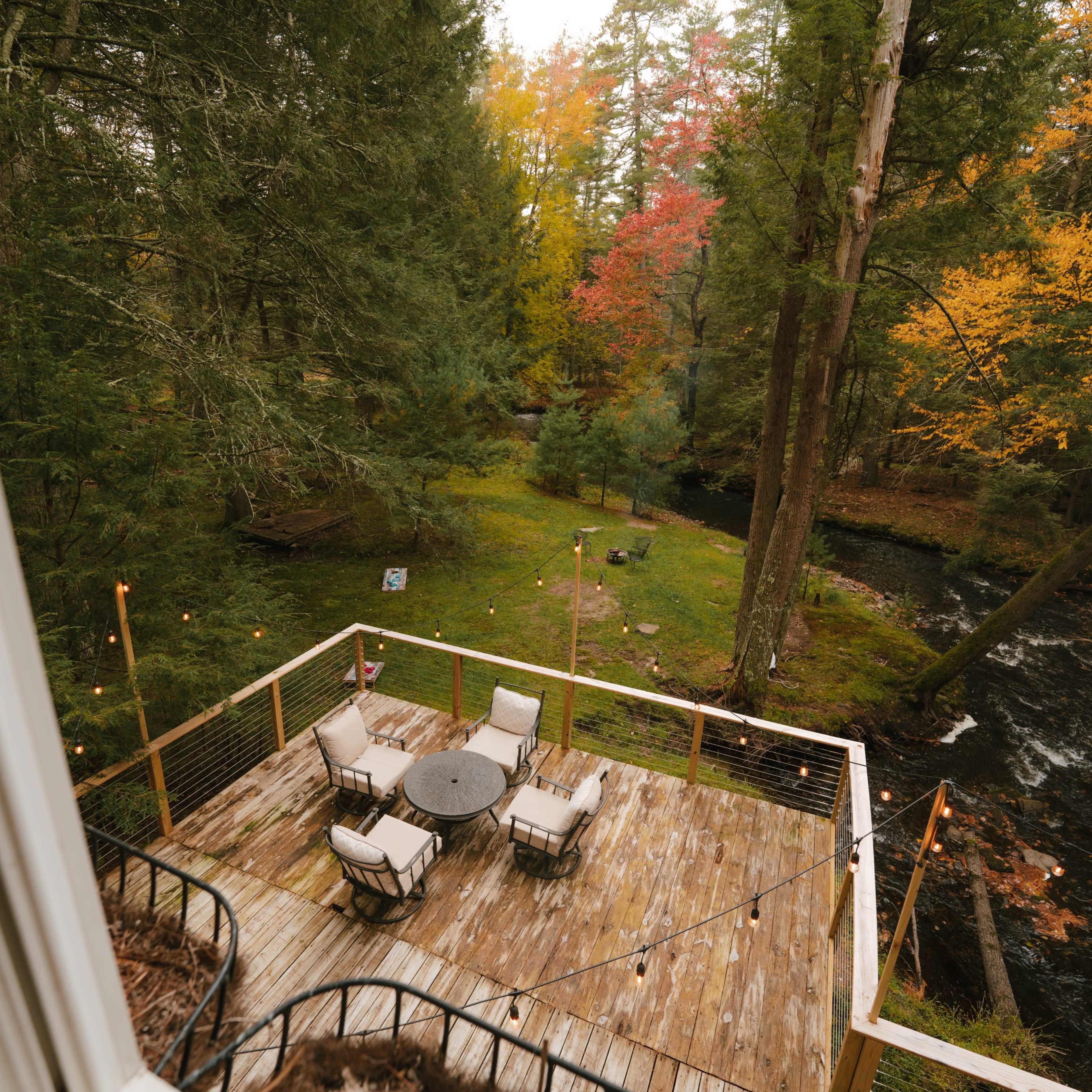 A wooden deck with chairs and a table overlooks a stream surrounded by trees with autumn foliage.