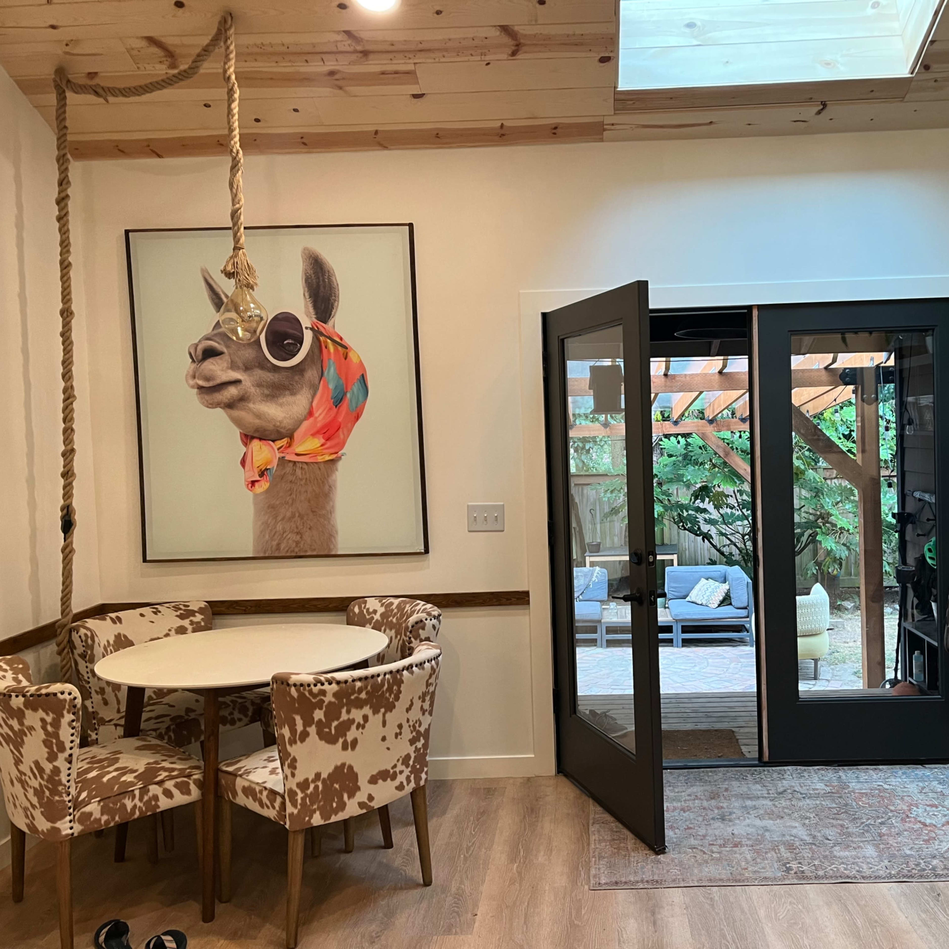 An entryway with a circular table and patterned chairs, a large framed portrait of a llama wearing sunglasses, and a pair of glass doors leading to a patio area.