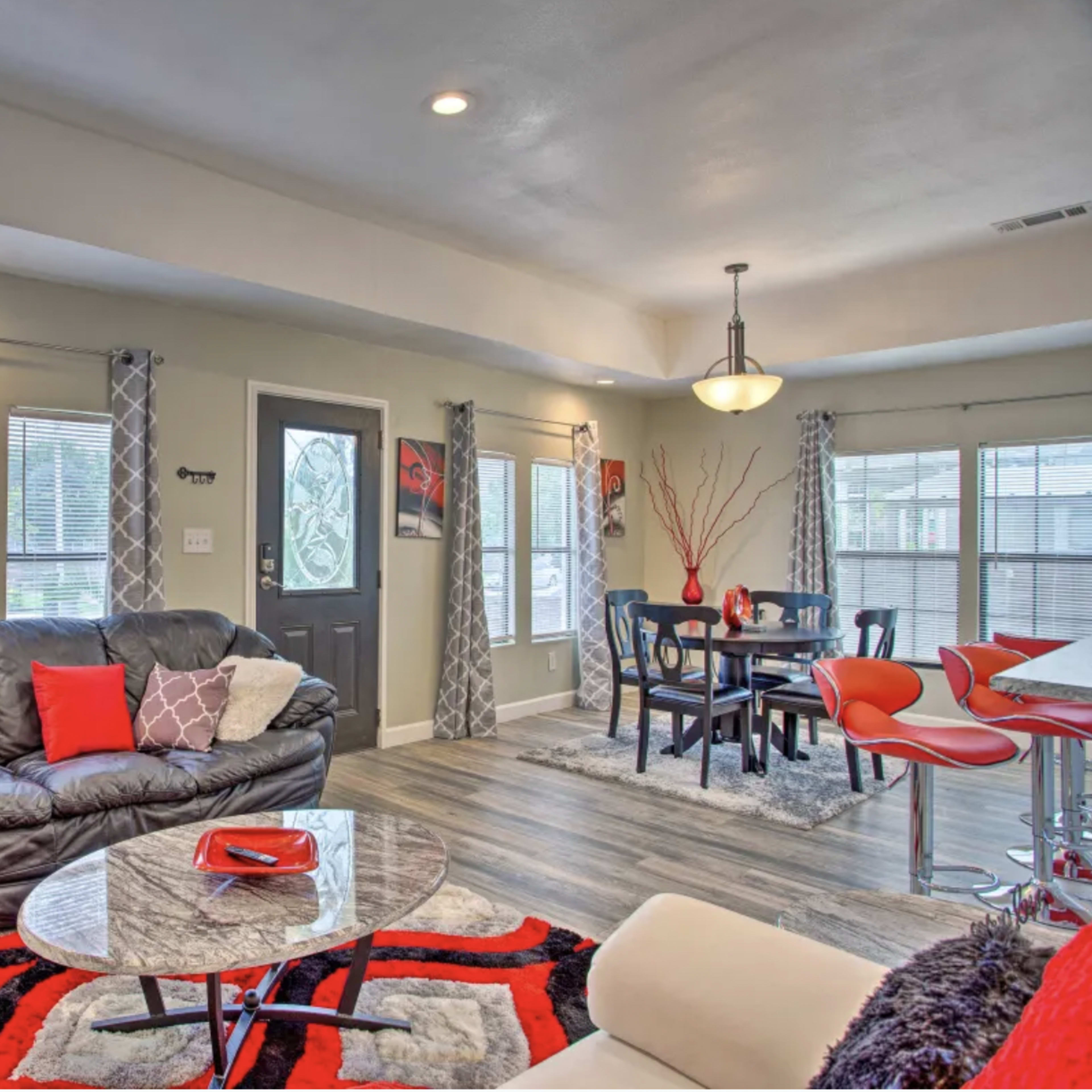 A modern living room and dining area with a black leather sofa, a round coffee table, and a dining table surrounded by red chairs, illuminated by natural light through multiple windows.