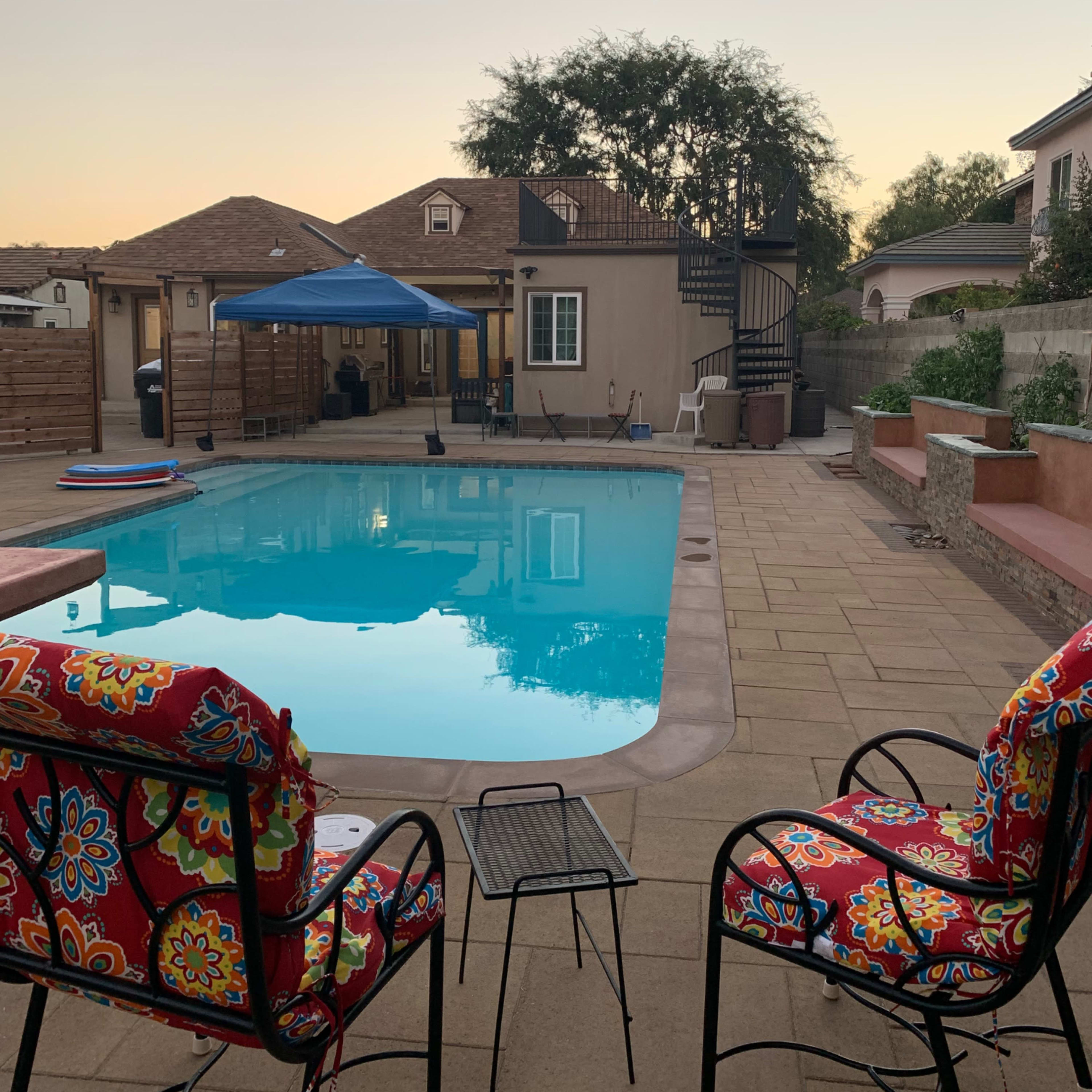 The scene shows a backyard with a swimming pool, two colorful chairs, and a home with a spiral staircase in the background during sunset.