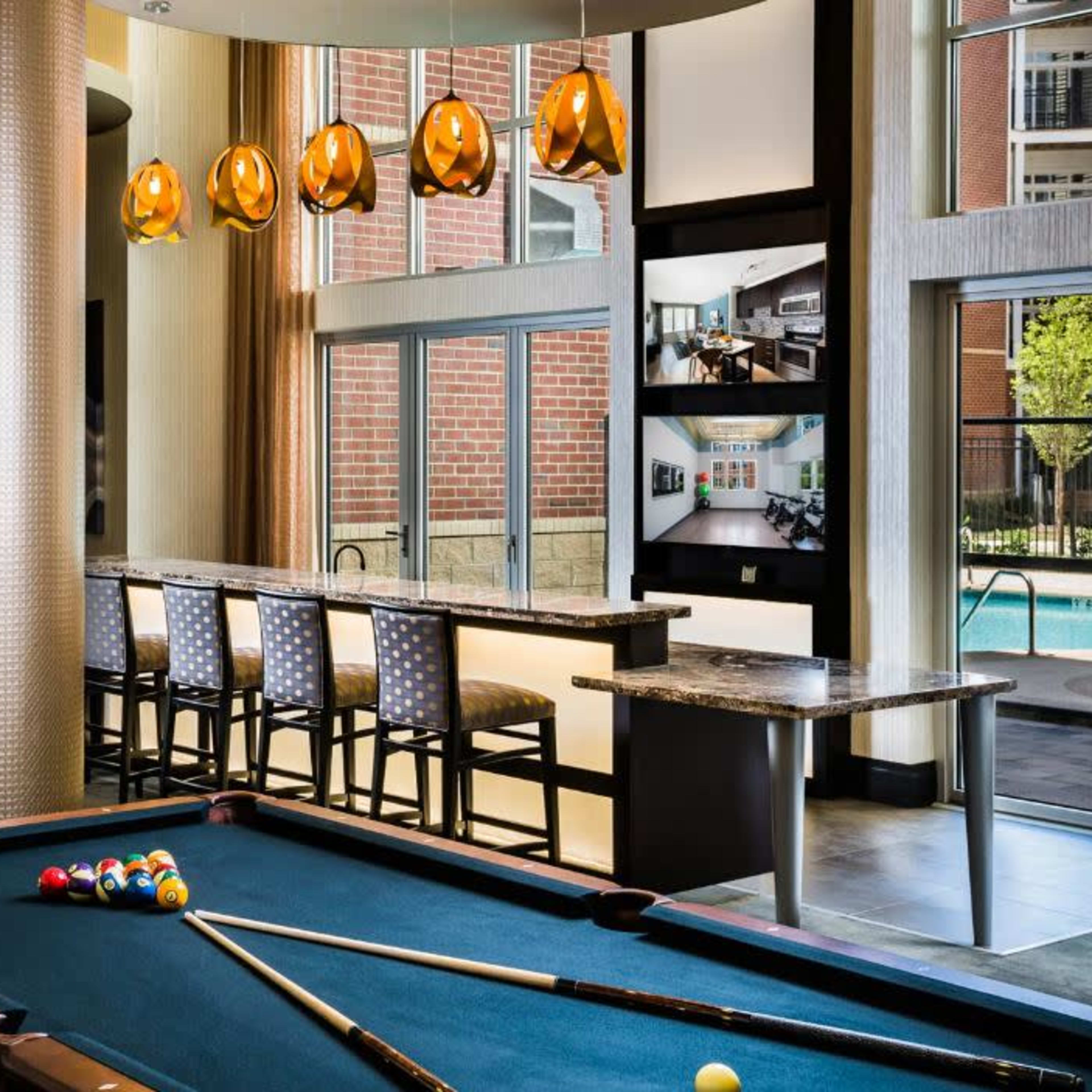 A modern lounge area with a pool table, a bar with stools, and large windows overlooking an outdoor pool.