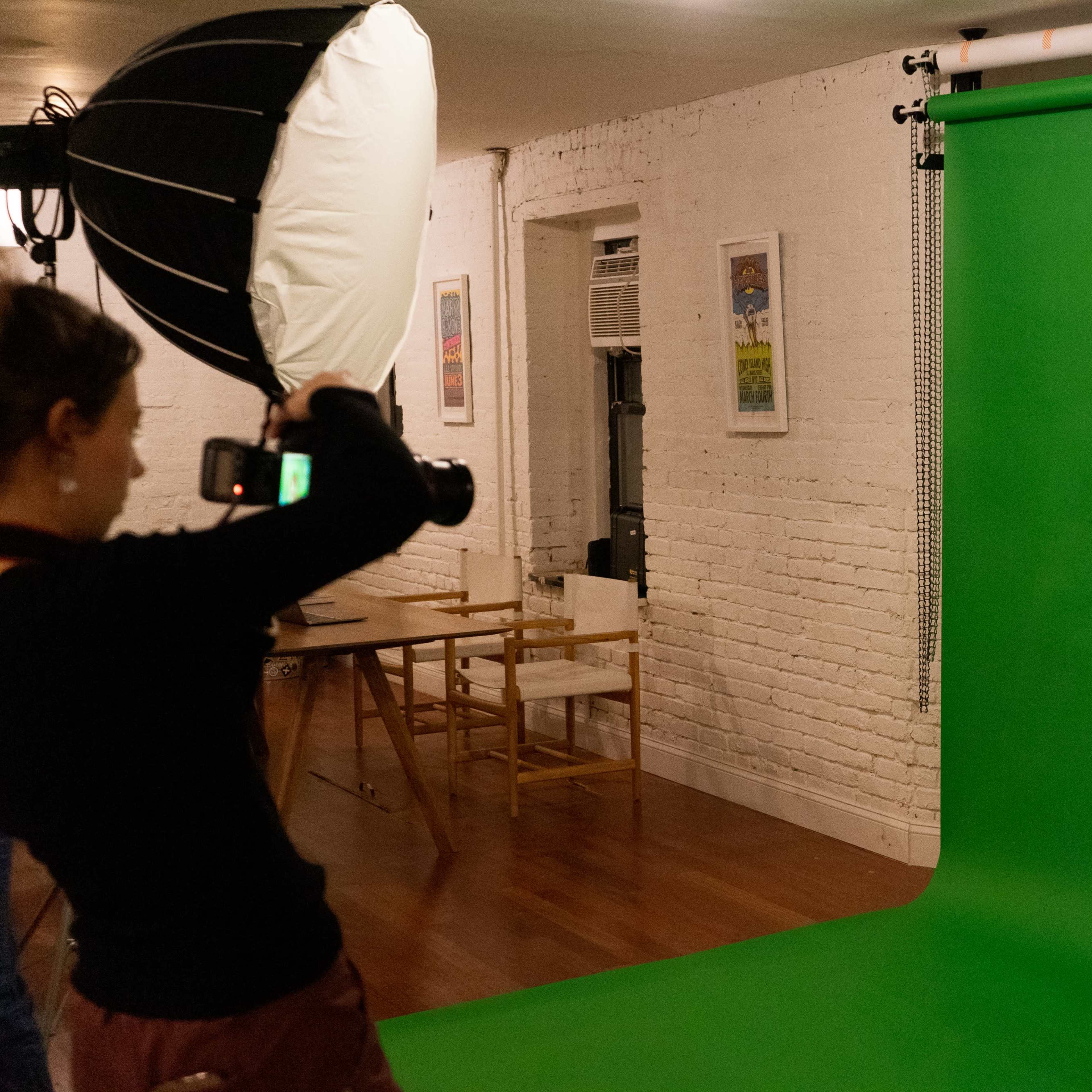 A model poses in front of a green screen while a photographer captures her image with a camera.