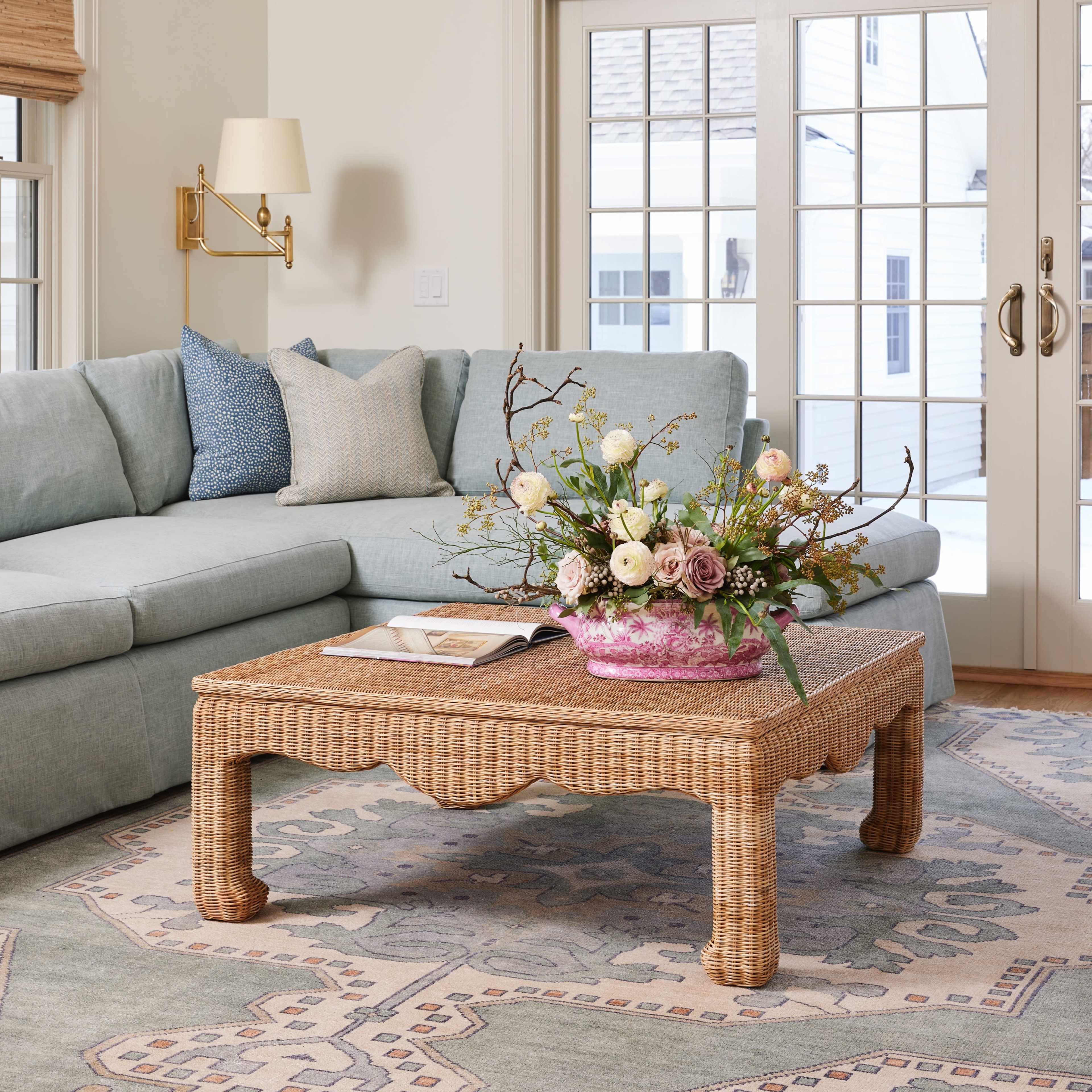 A light-filled living room features a pale blue sectional sofa, a woven coffee table with a floral arrangement, and large glass doors leading outside.