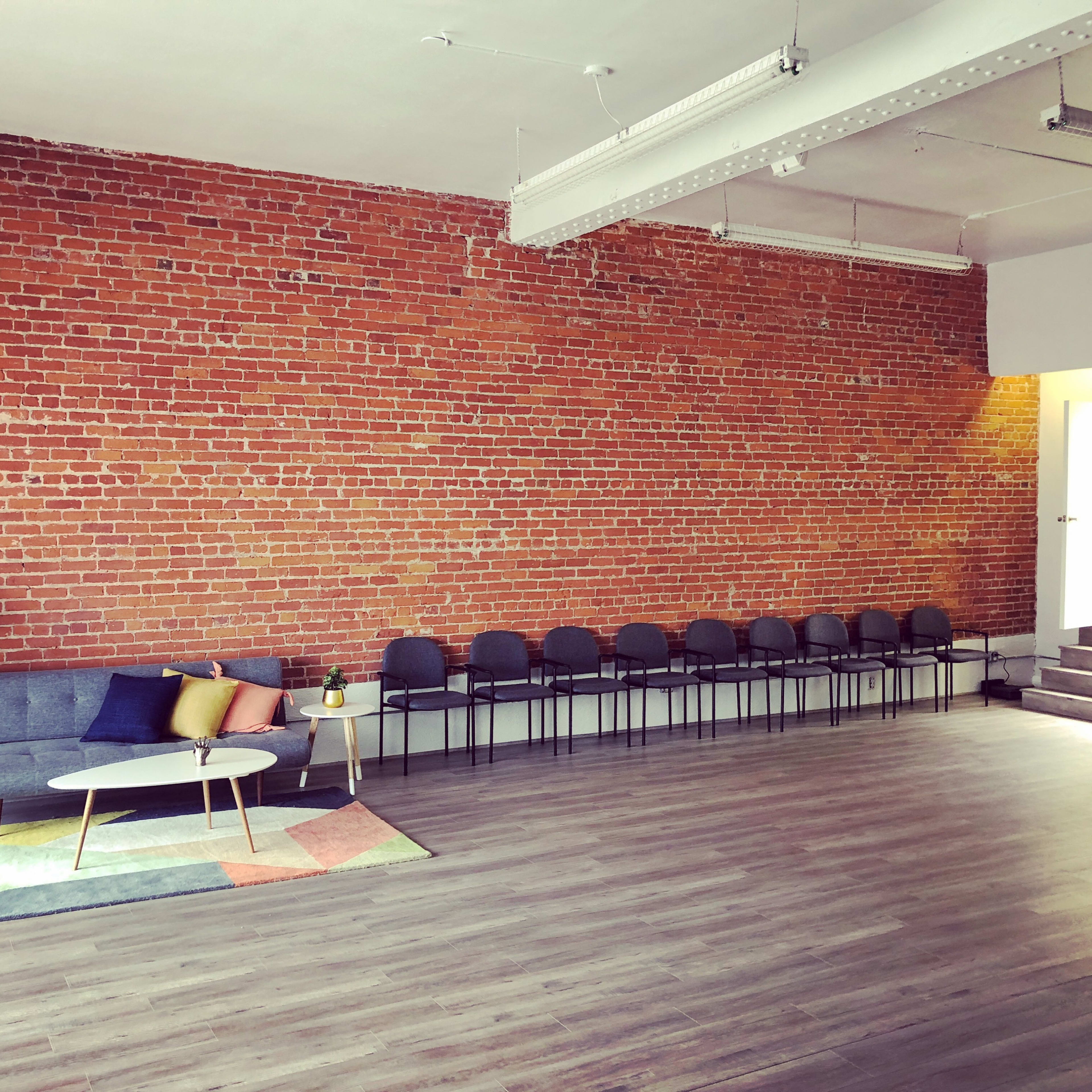 A spacious room with a brick wall, a gray sofa with colorful pillows, a patterned rug, and a row of chairs along one side.