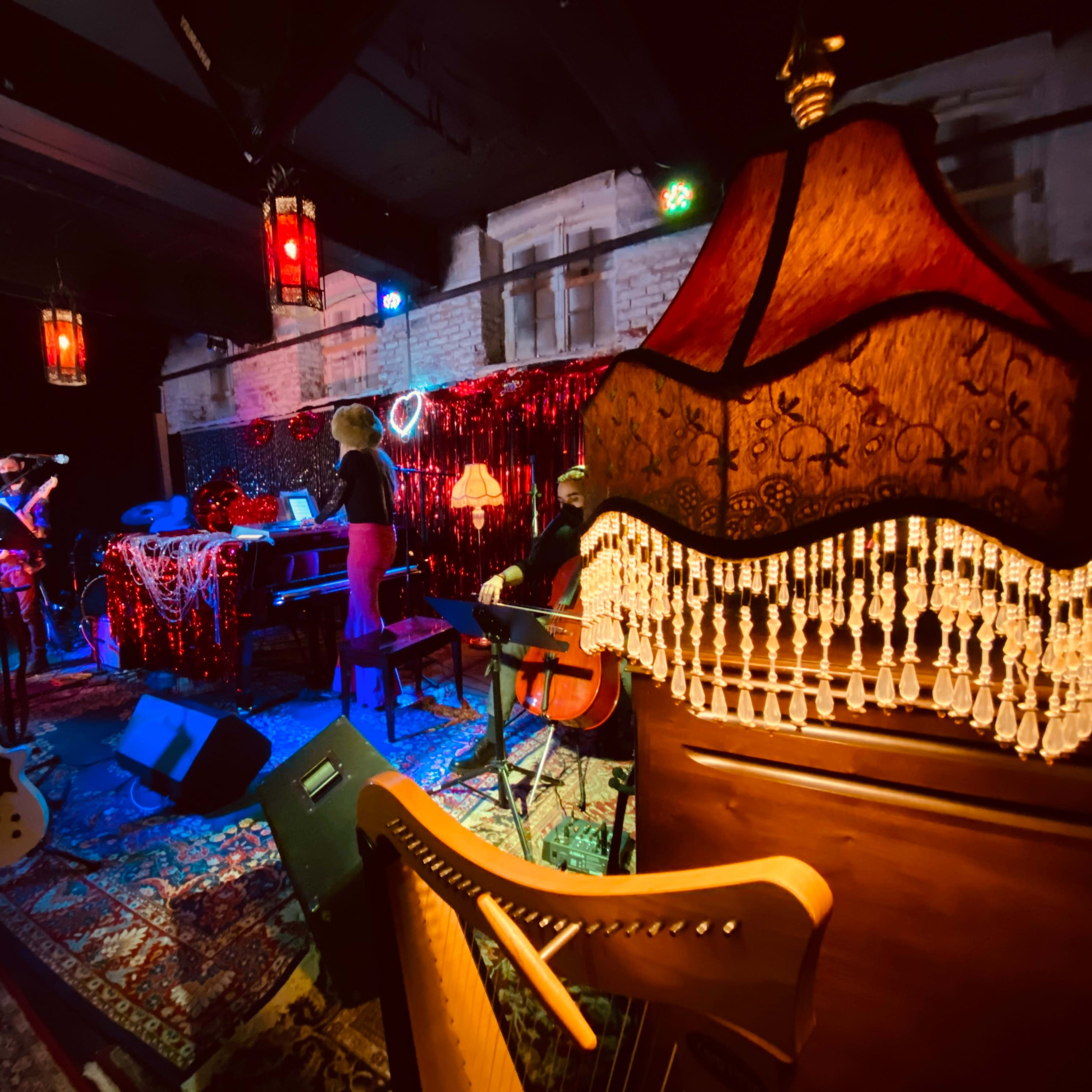 A dimly lit music venue with musicians on stage, surrounded by decorative elements like lamps and a harp.