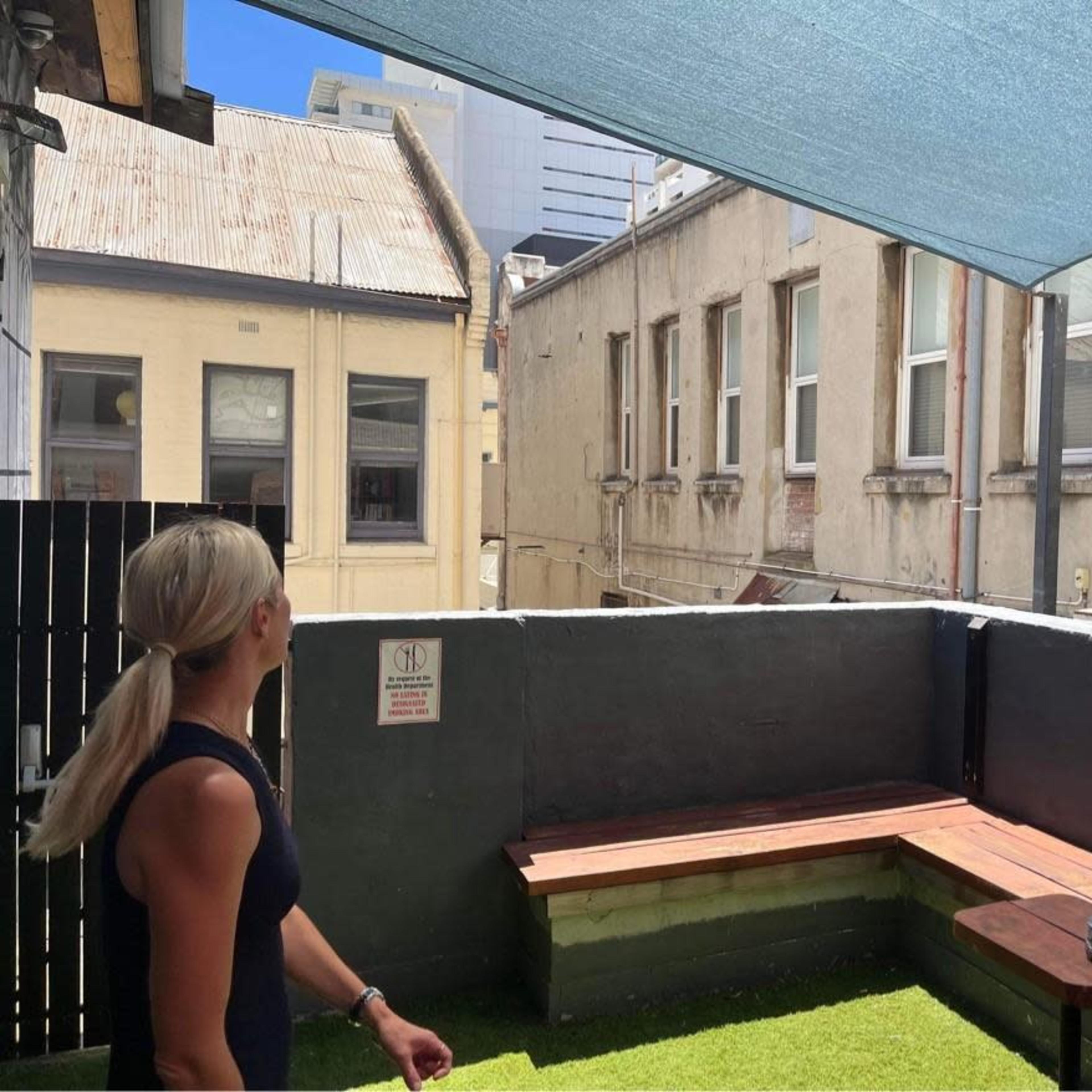 A woman stands on a rooftop terrace overlooking brick buildings and a blue sky, with a shaded area and seating made of wooden benches.