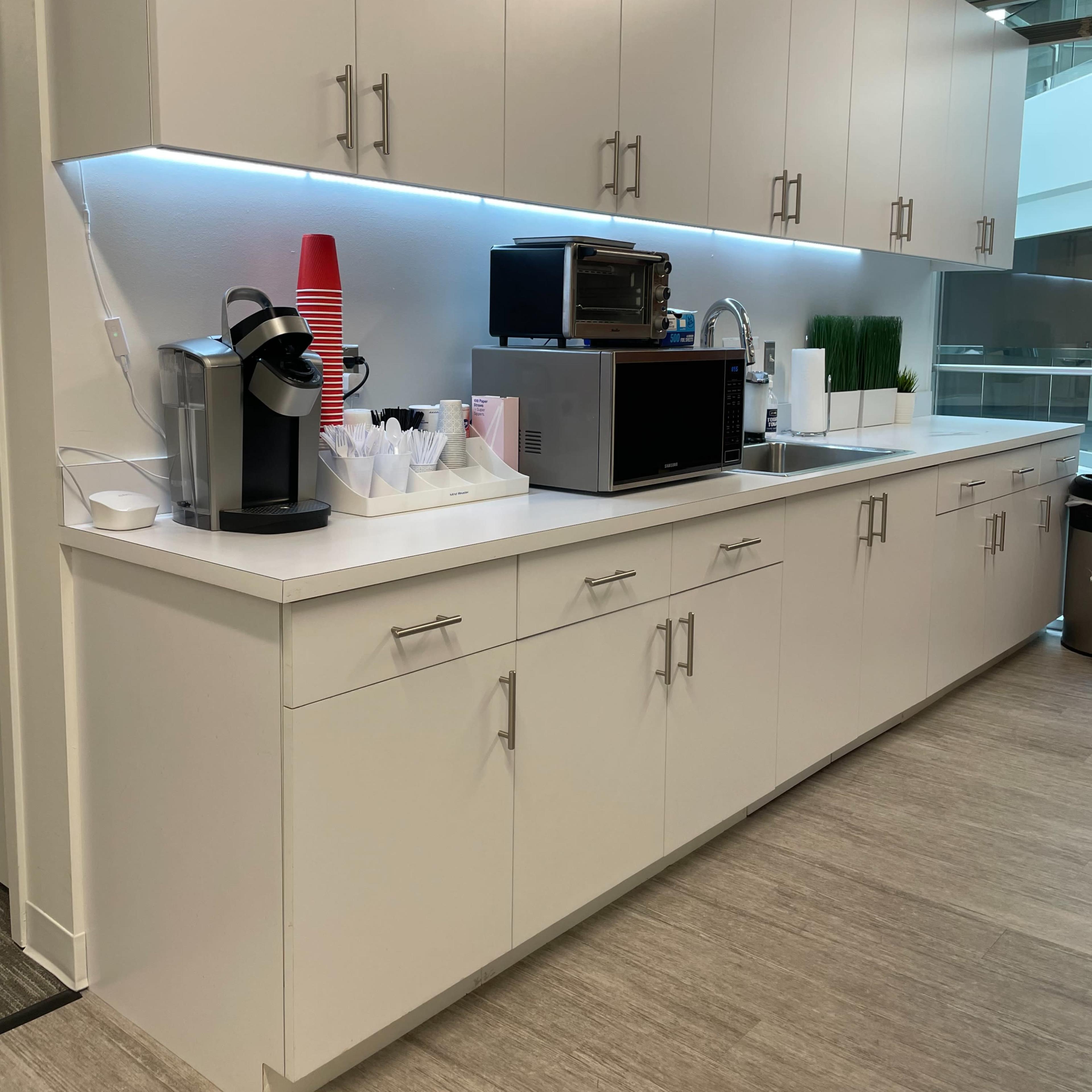 A modern kitchen area featuring white cabinets, a microwave, a coffee maker, stacked cups, and decorative greenery on a countertop.