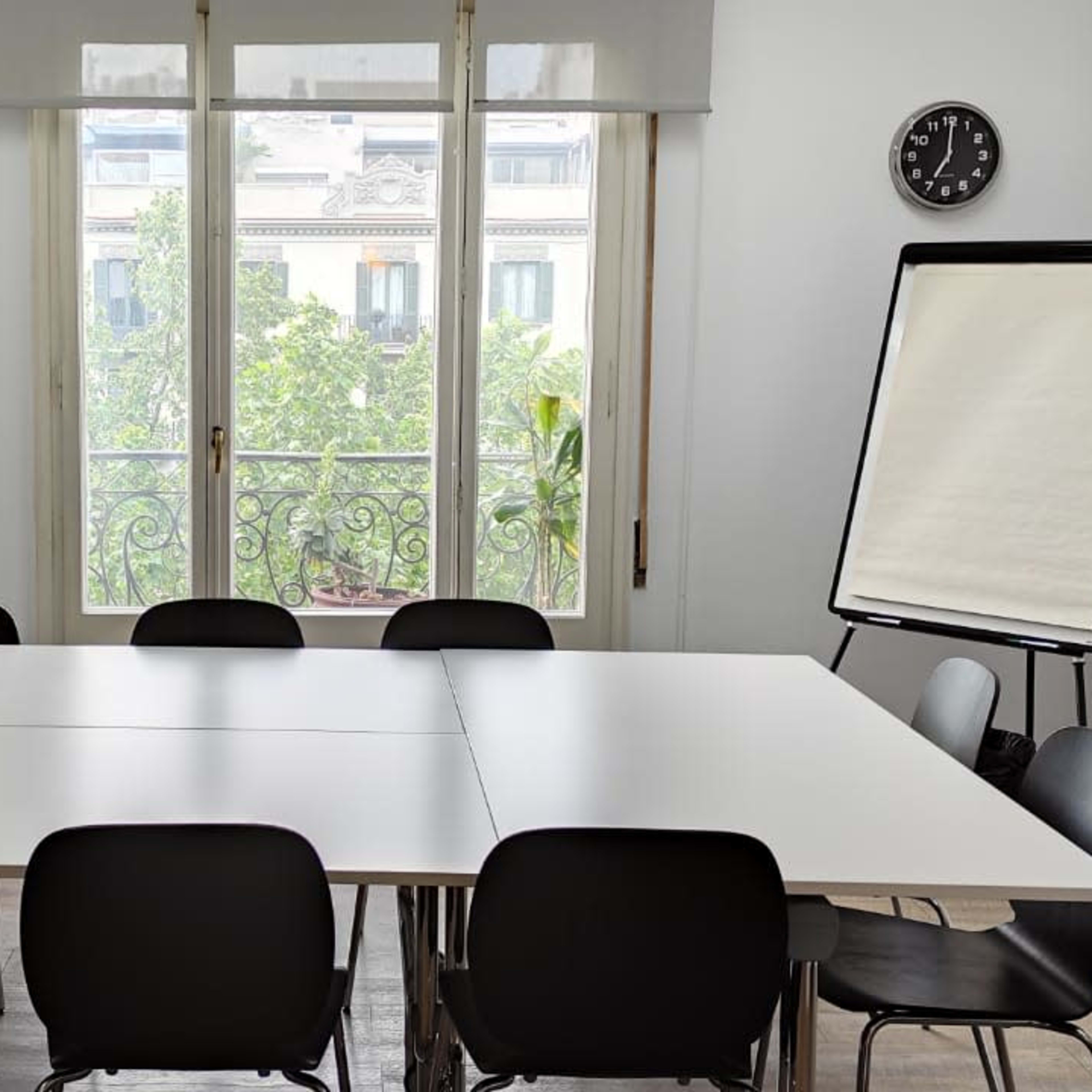 A modern meeting room with a large table surrounded by black chairs, a flip chart, a clock on the wall, and a window overlooking a green view.