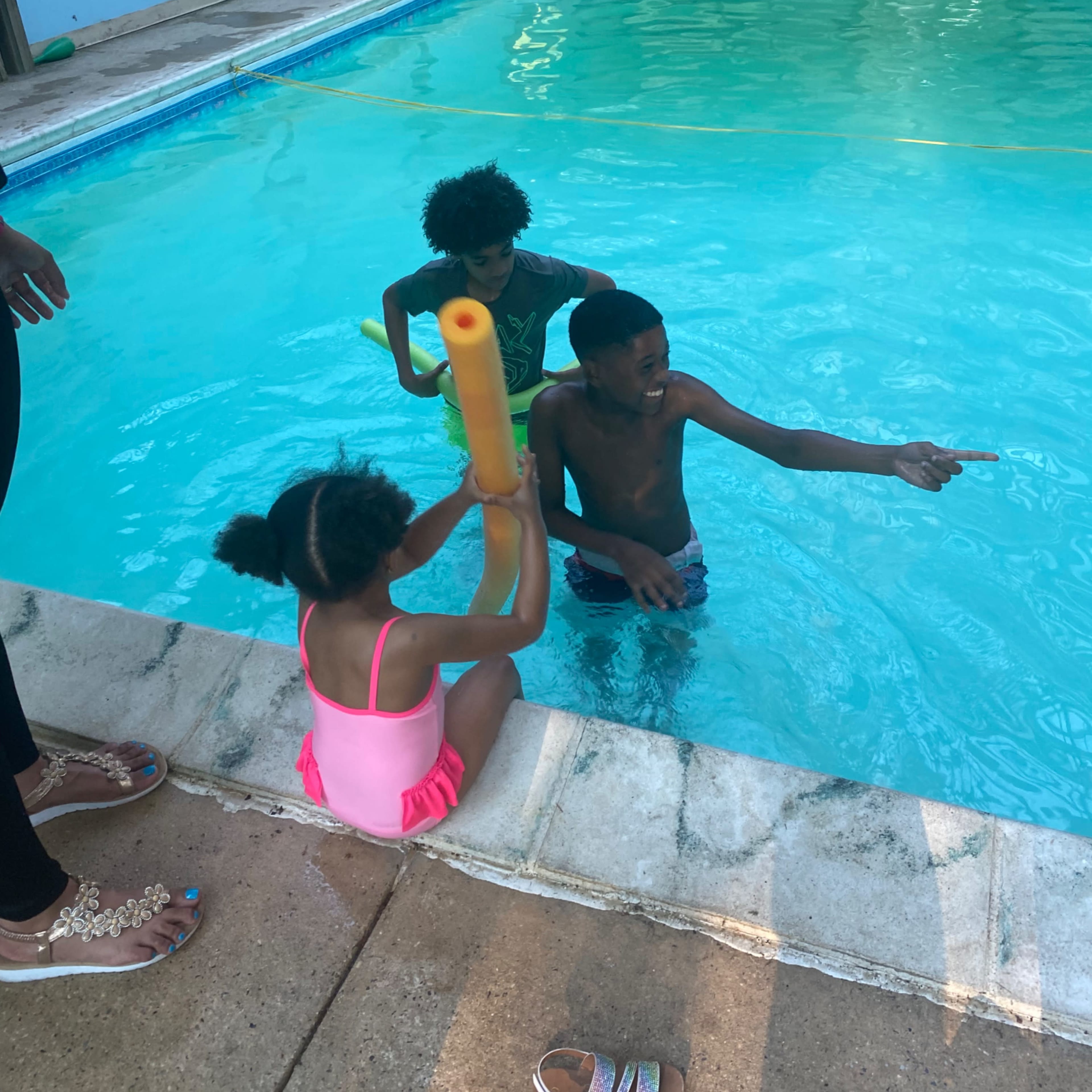 Three children are playing in and around a swimming pool, with two in the water and one sitting on the edge.