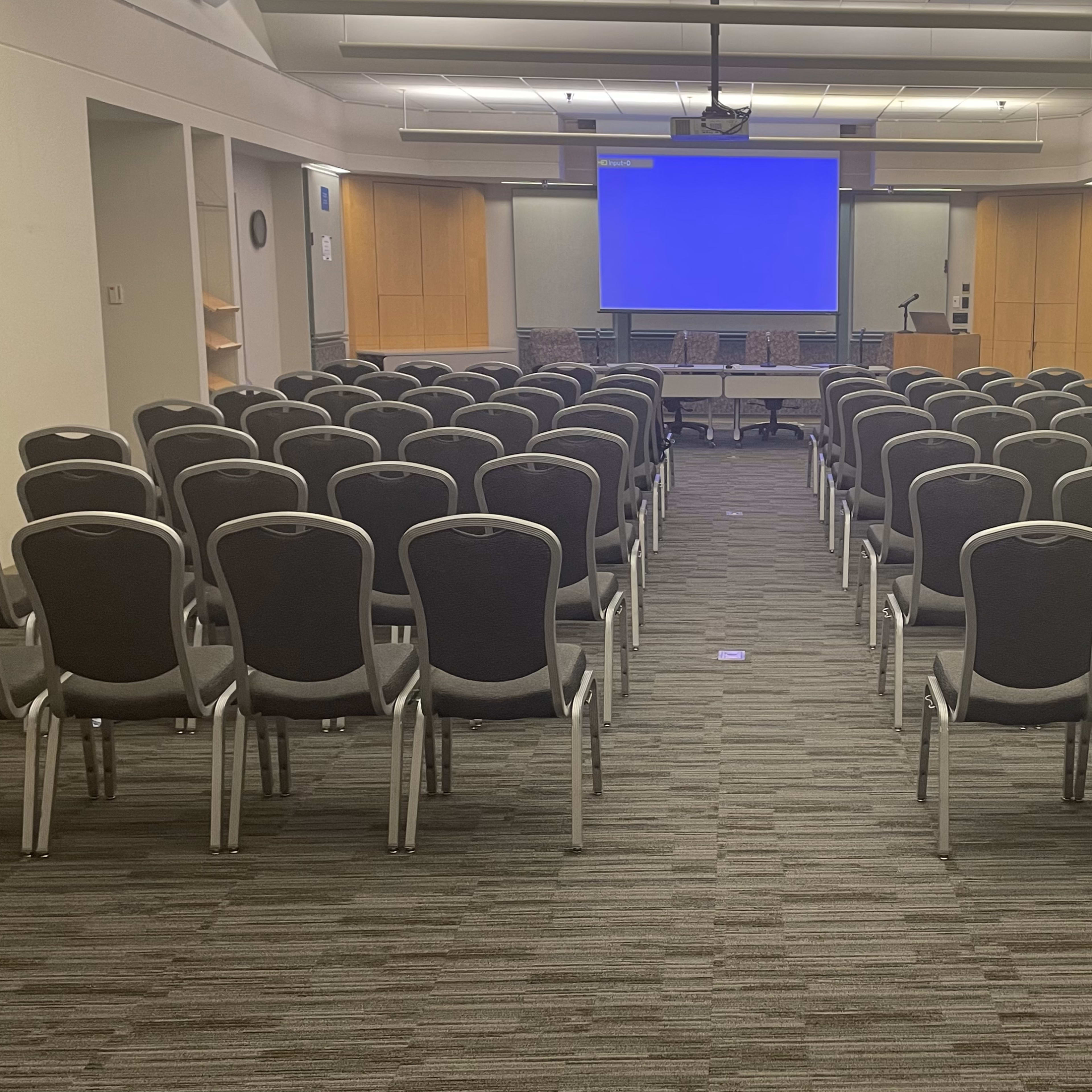A conference room with rows of black chairs facing a large blank screen.