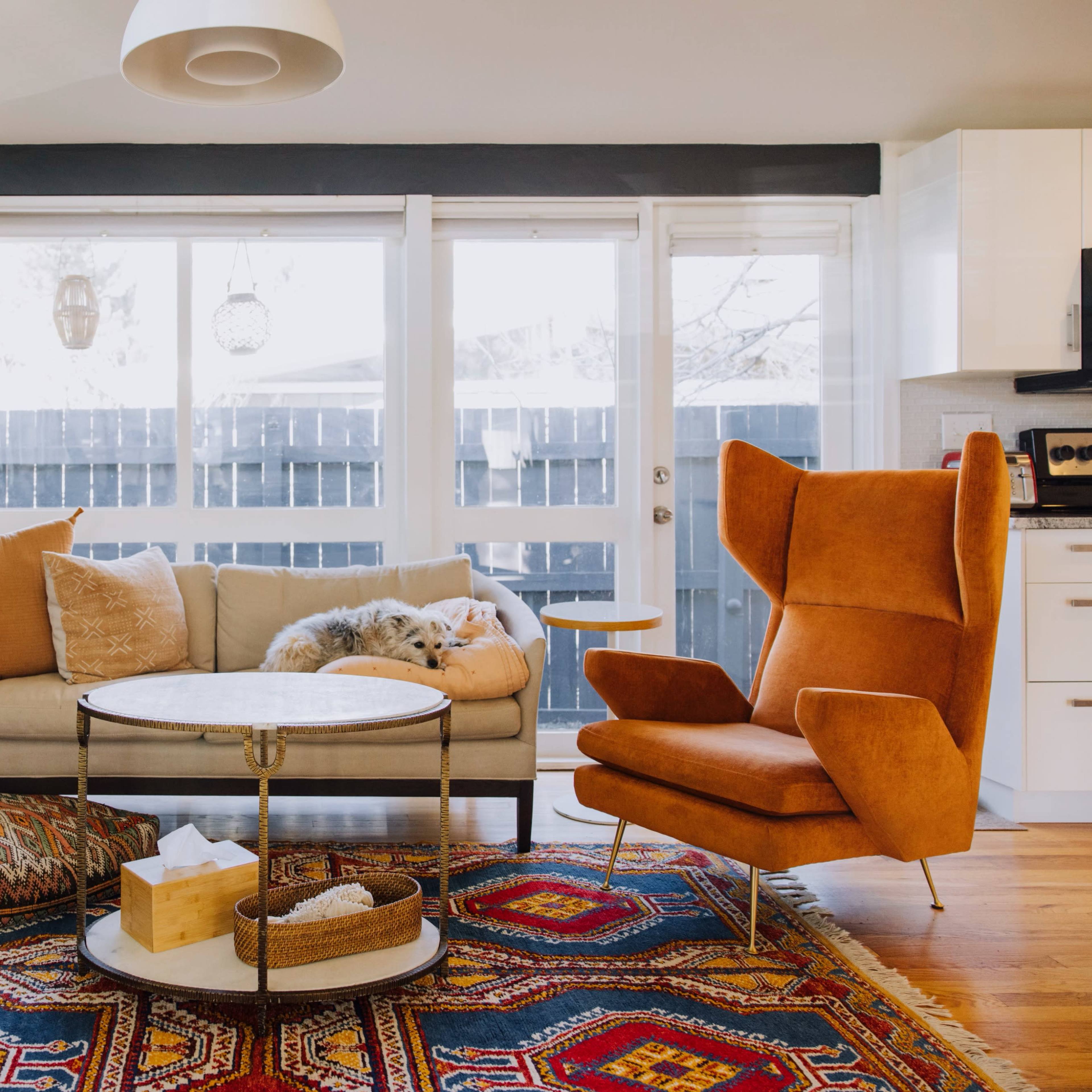 A cozy living room features a beige sofa, an orange accent chair, and a round coffee table, all set against a backdrop of large windows and a vibrant area rug.
