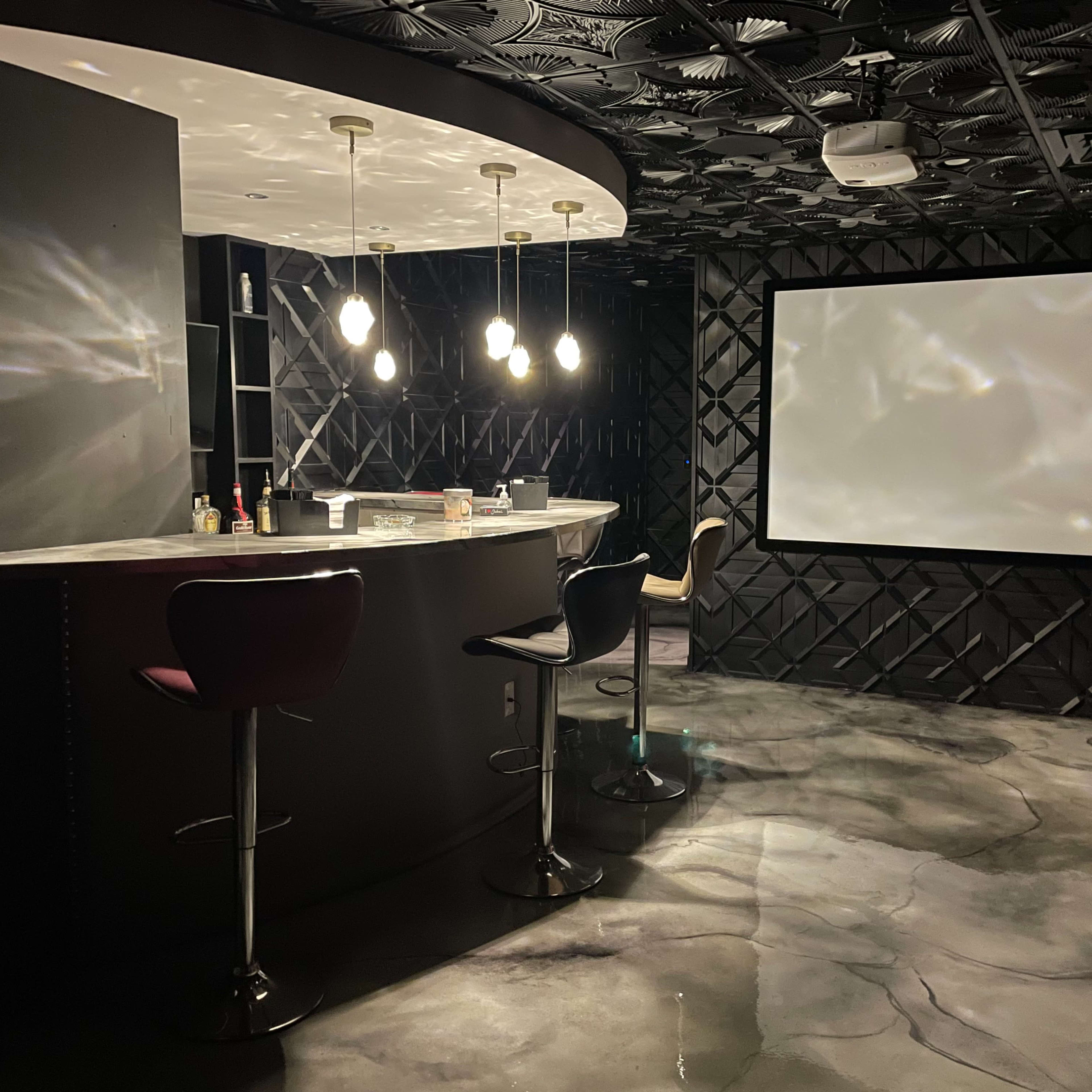 A stylish room with a bar area featuring modern stools and a large blank screen on one wall, illuminated by pendant lights.