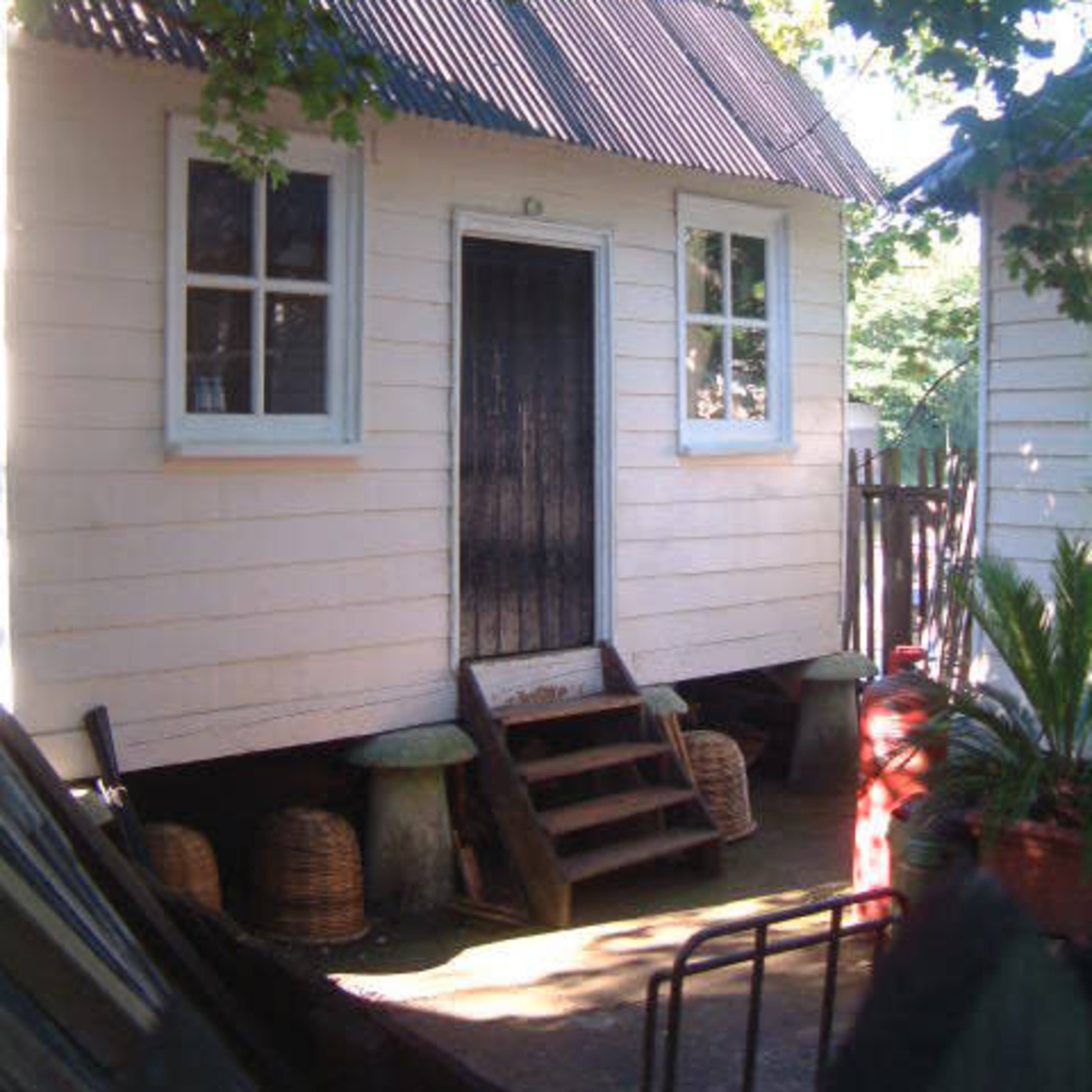A small white wooden house with a dark door and a sloped metal roof, surrounded by trees and outdoor furnishings.