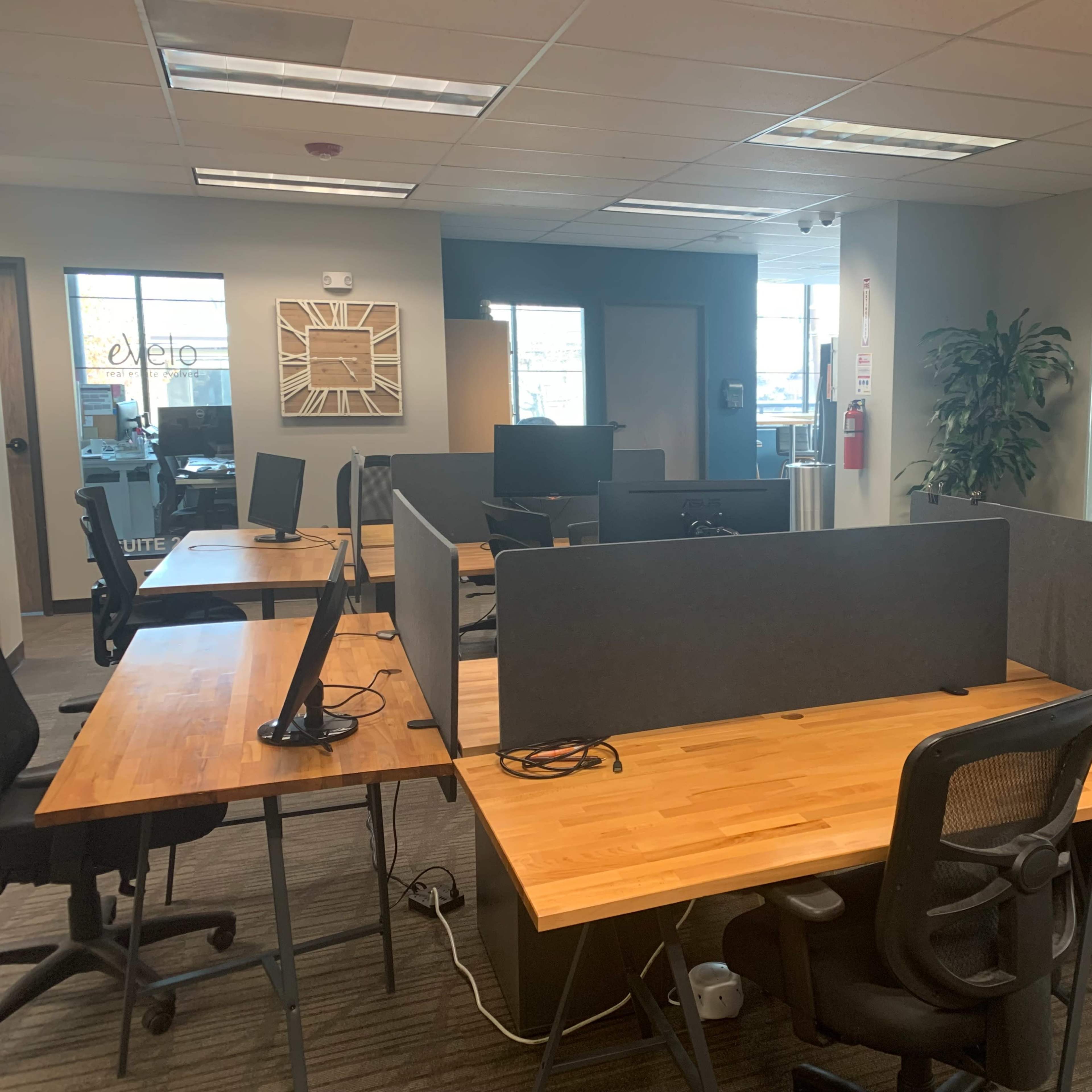 An office space with several workstations featuring wooden desks and ergonomic chairs, separated by gray partition walls.