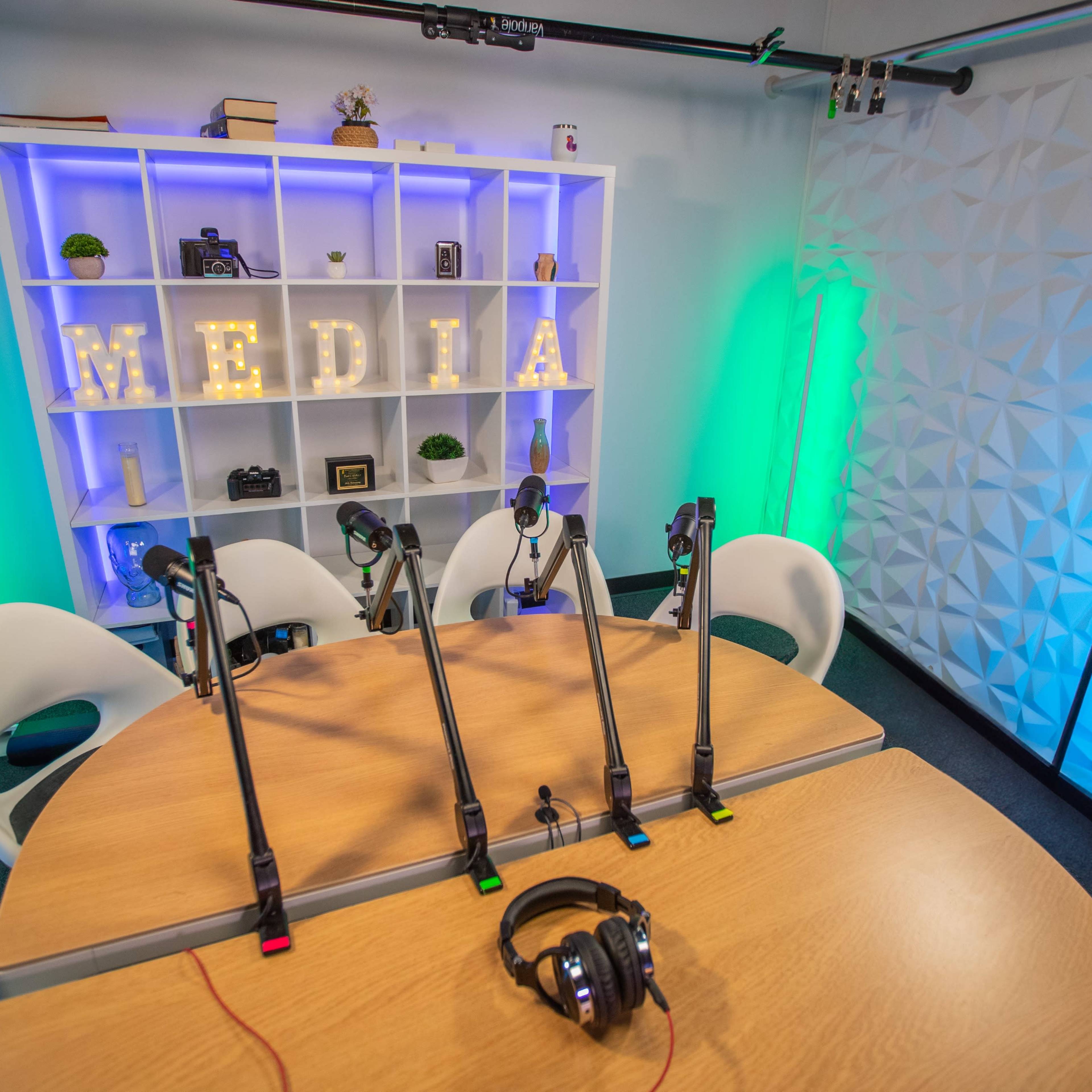 A recording studio is set up with a table, several microphones on stands, and chairs, alongside a brightly lit shelf displaying the word "MEDIA."