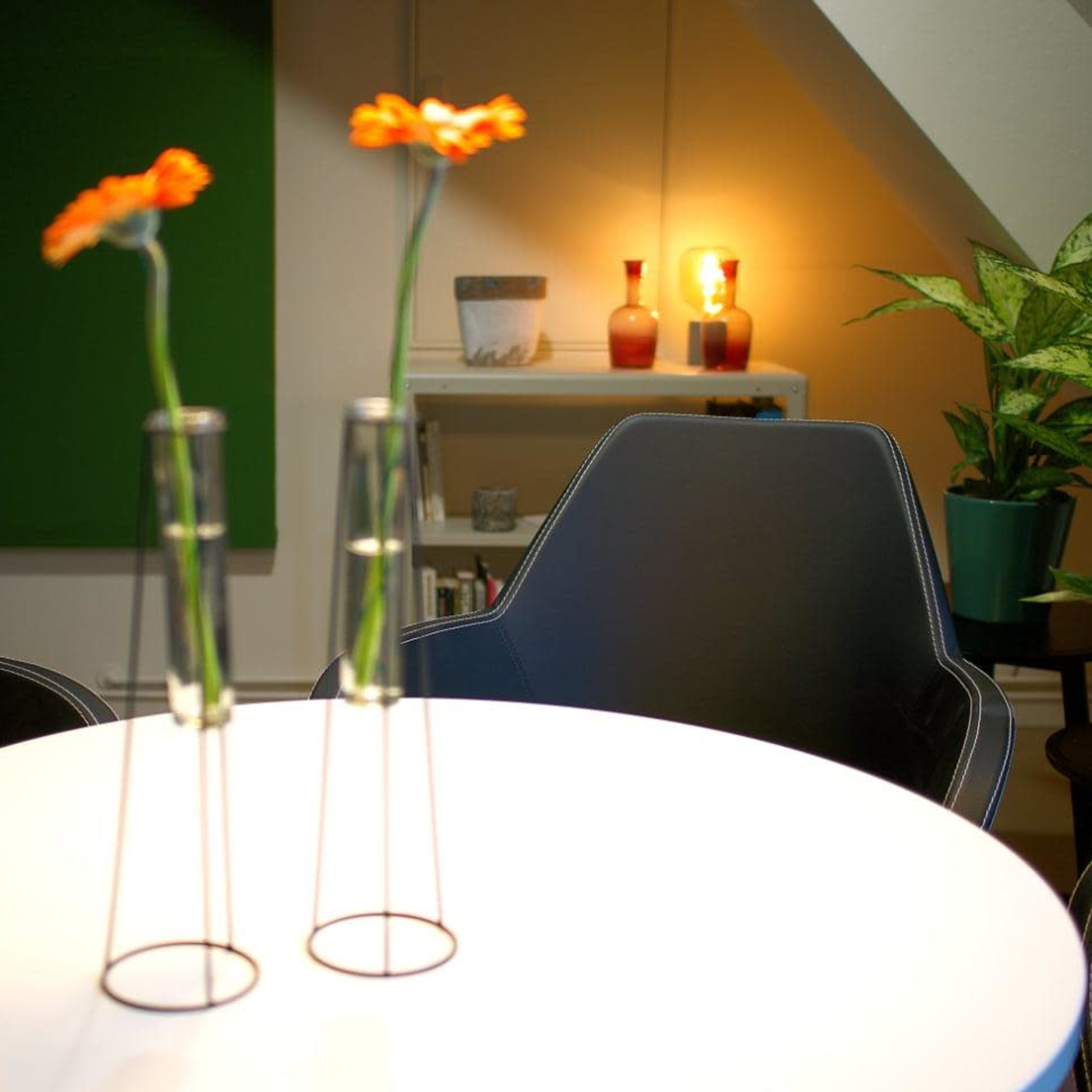 A round table with two test tube vases holding orange flowers is set in a modern room with a green wall and warm lighting.