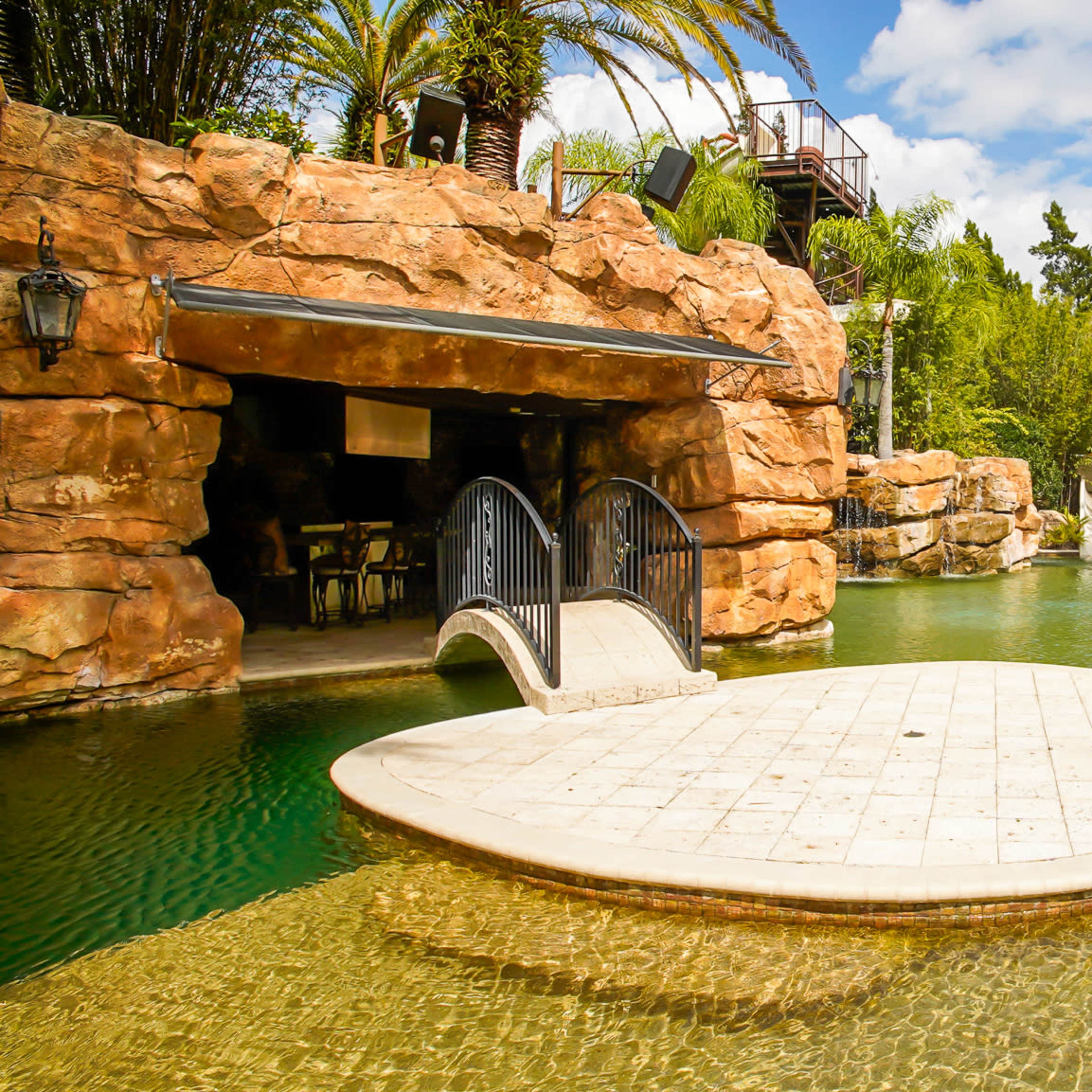 A landscaped outdoor space featuring a circular stone platform connected by a small bridge to a rocky cave-like structure, surrounded by palm trees and water.