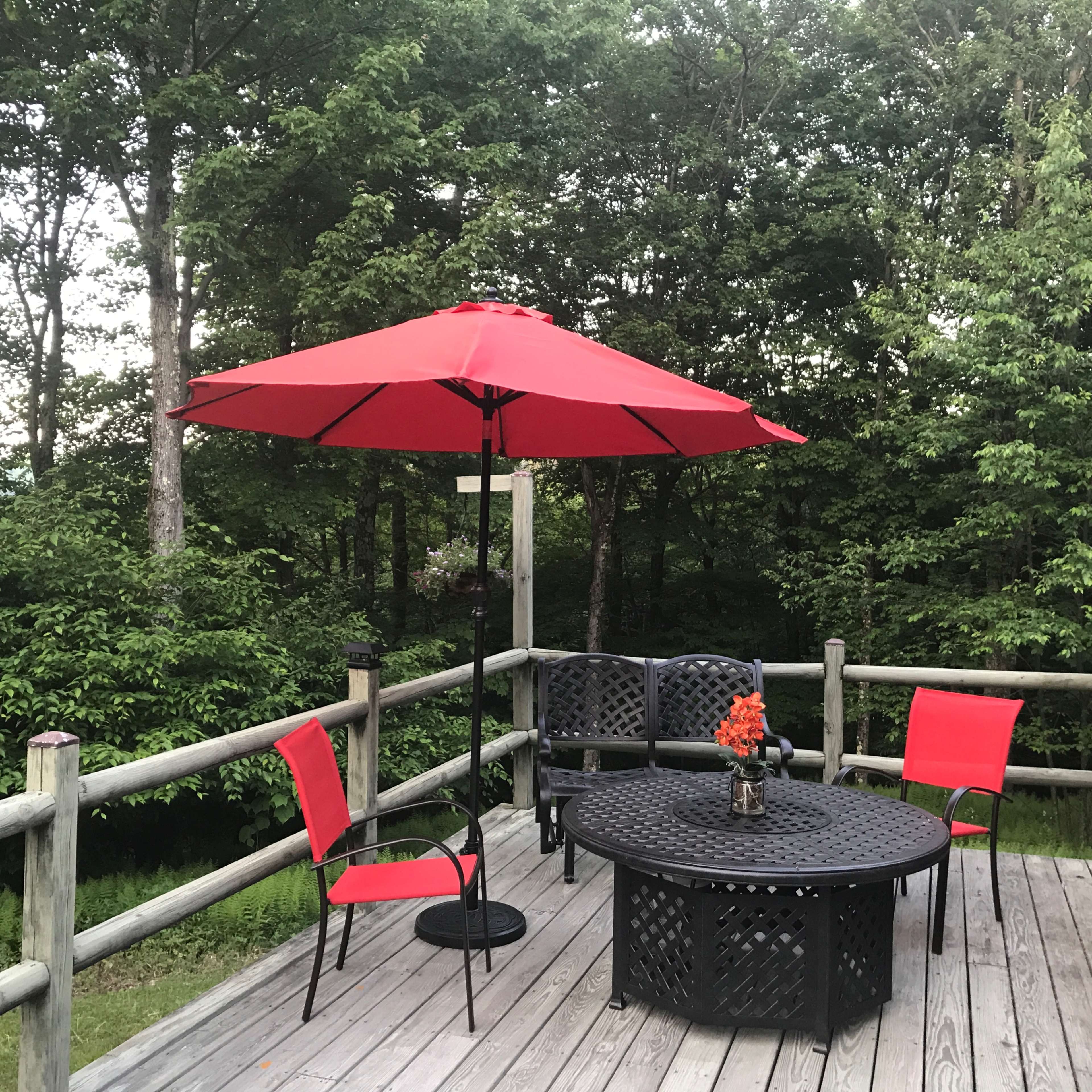 A red umbrella shades a circular black table surrounded by chairs on a wooden deck overlooking greenery.