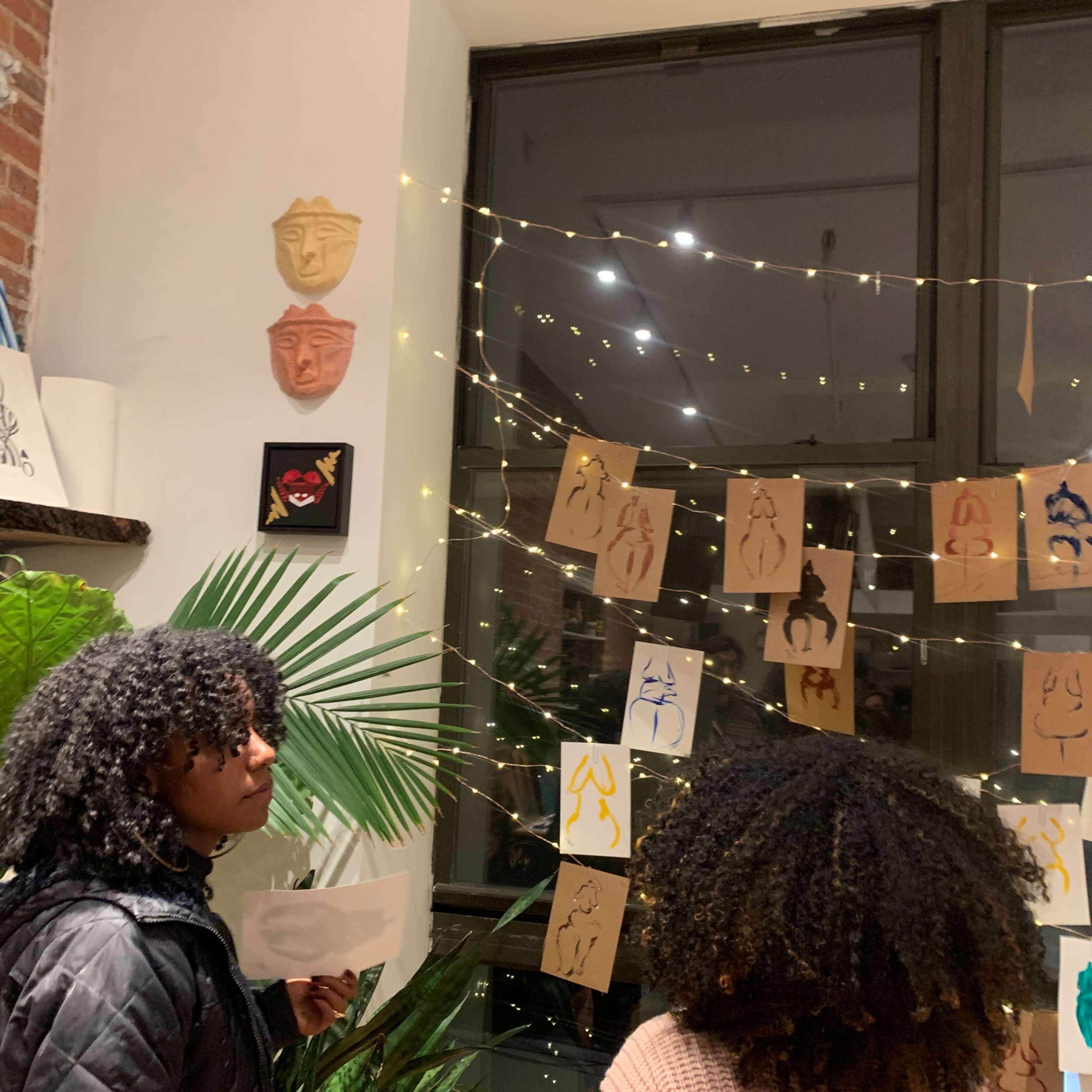 Two individuals observe artwork displayed on brown paper hanging near a window adorned with string lights in a cozy indoor setting.