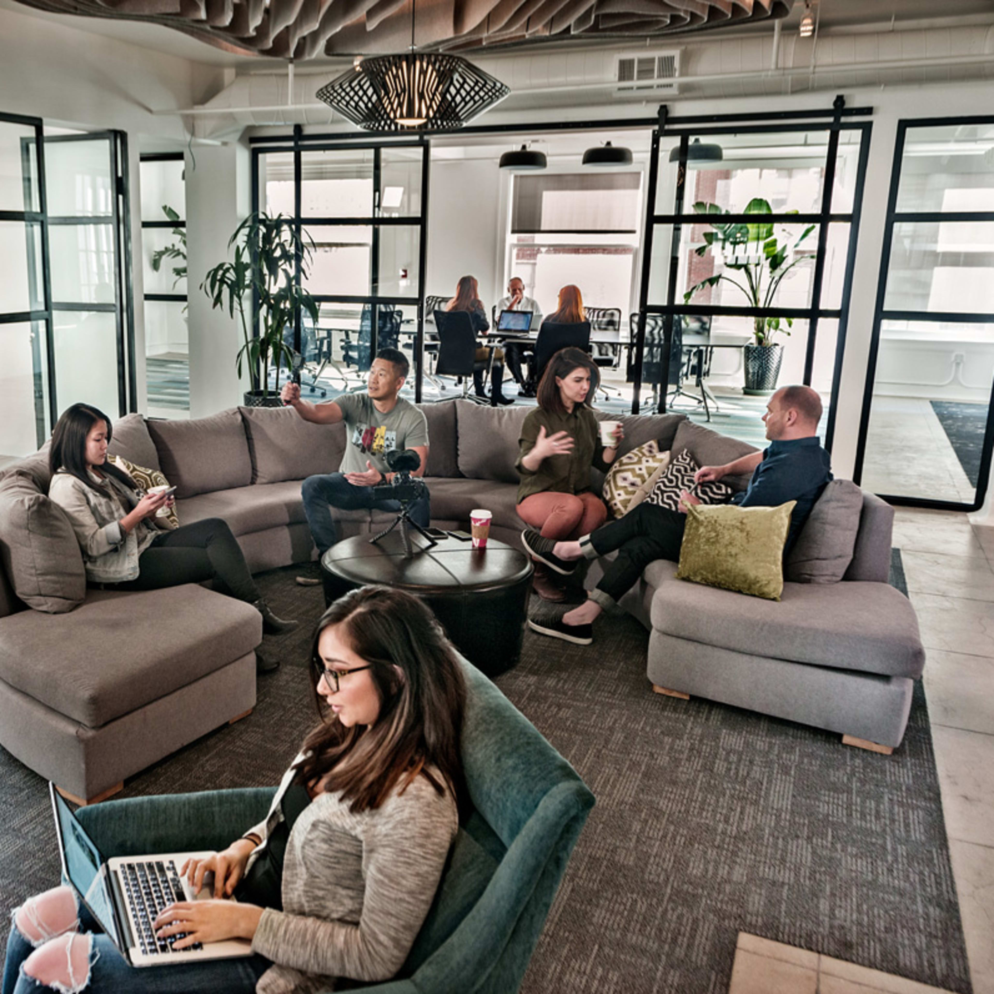 A group of people are working and engaging in conversation in a modern office space with a lounge area and glass meeting rooms.