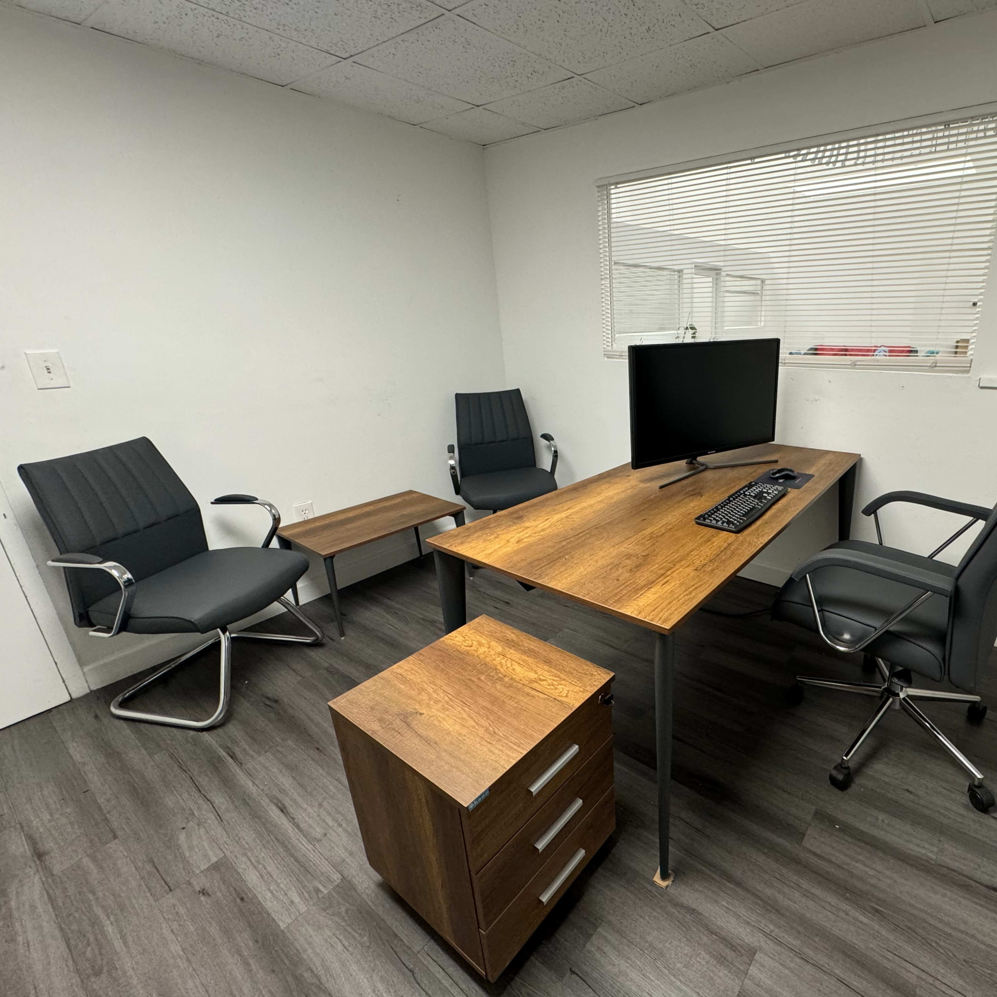 A minimalist office space with a wooden desk, a computer monitor, three chairs, a side table, and a storage unit.