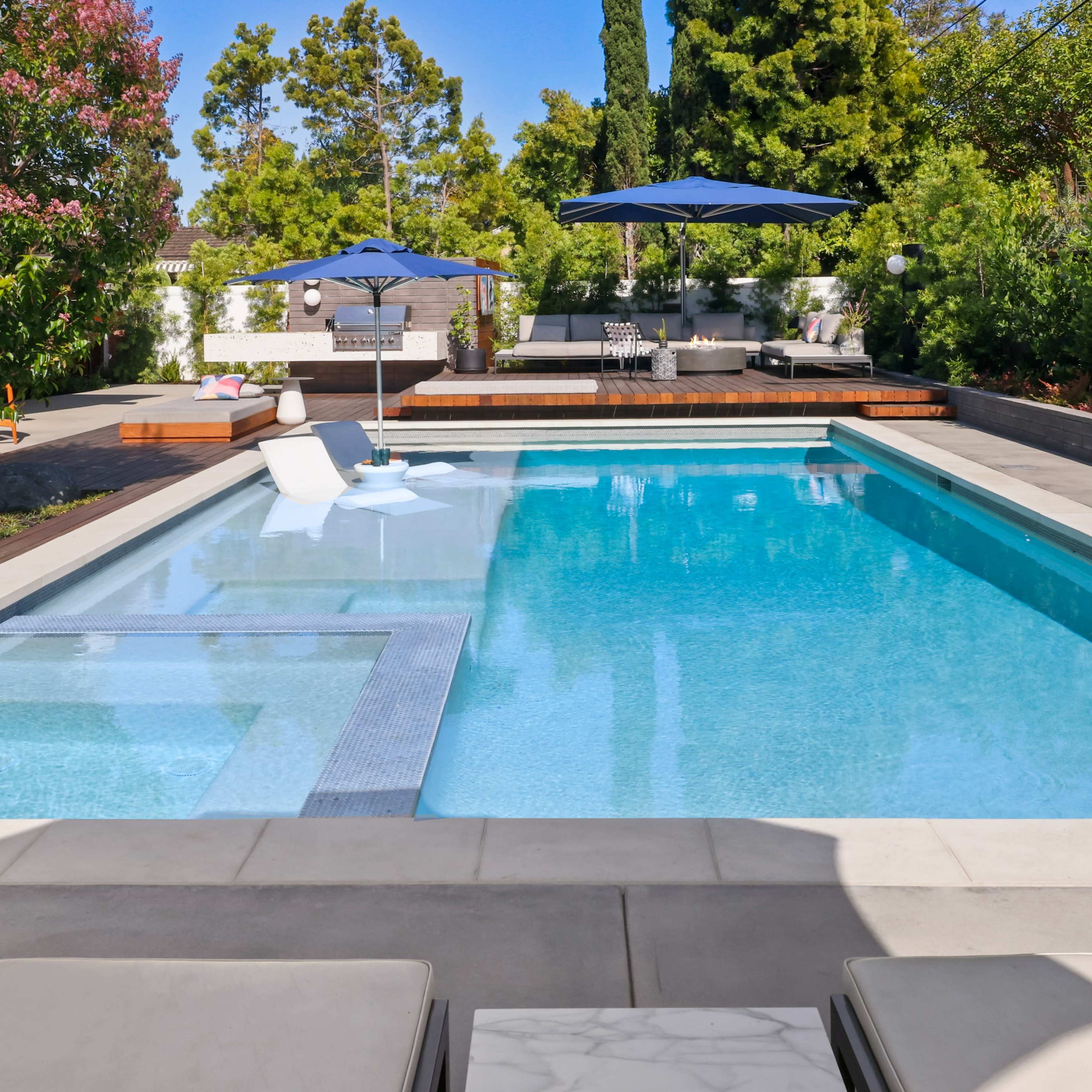 A clear swimming pool surrounded by a patio, lounge chairs, and shaded seating areas in a landscaped backyard.