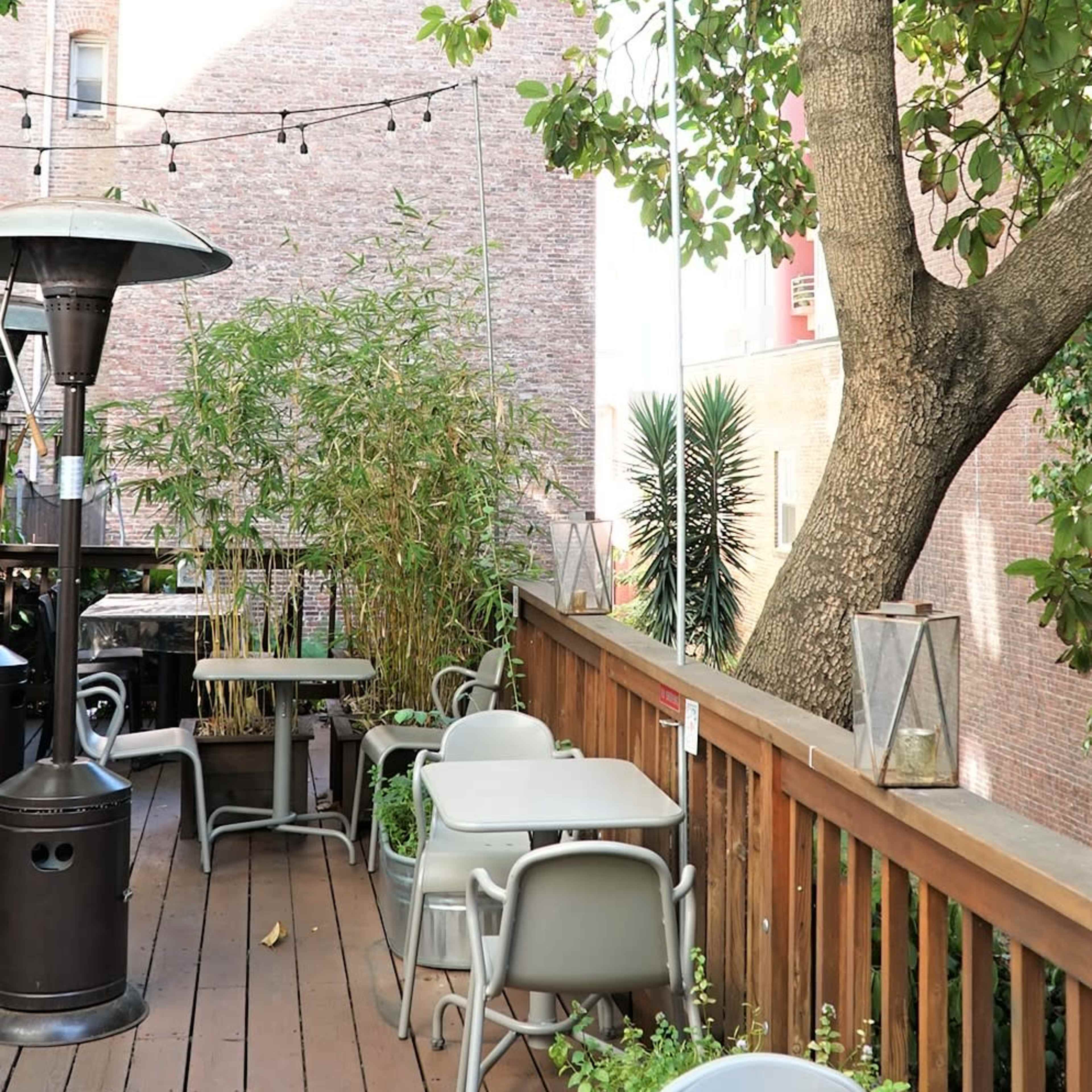 A wooden deck terrace with metal tables and chairs, surrounded by greenery and overhead string lights.