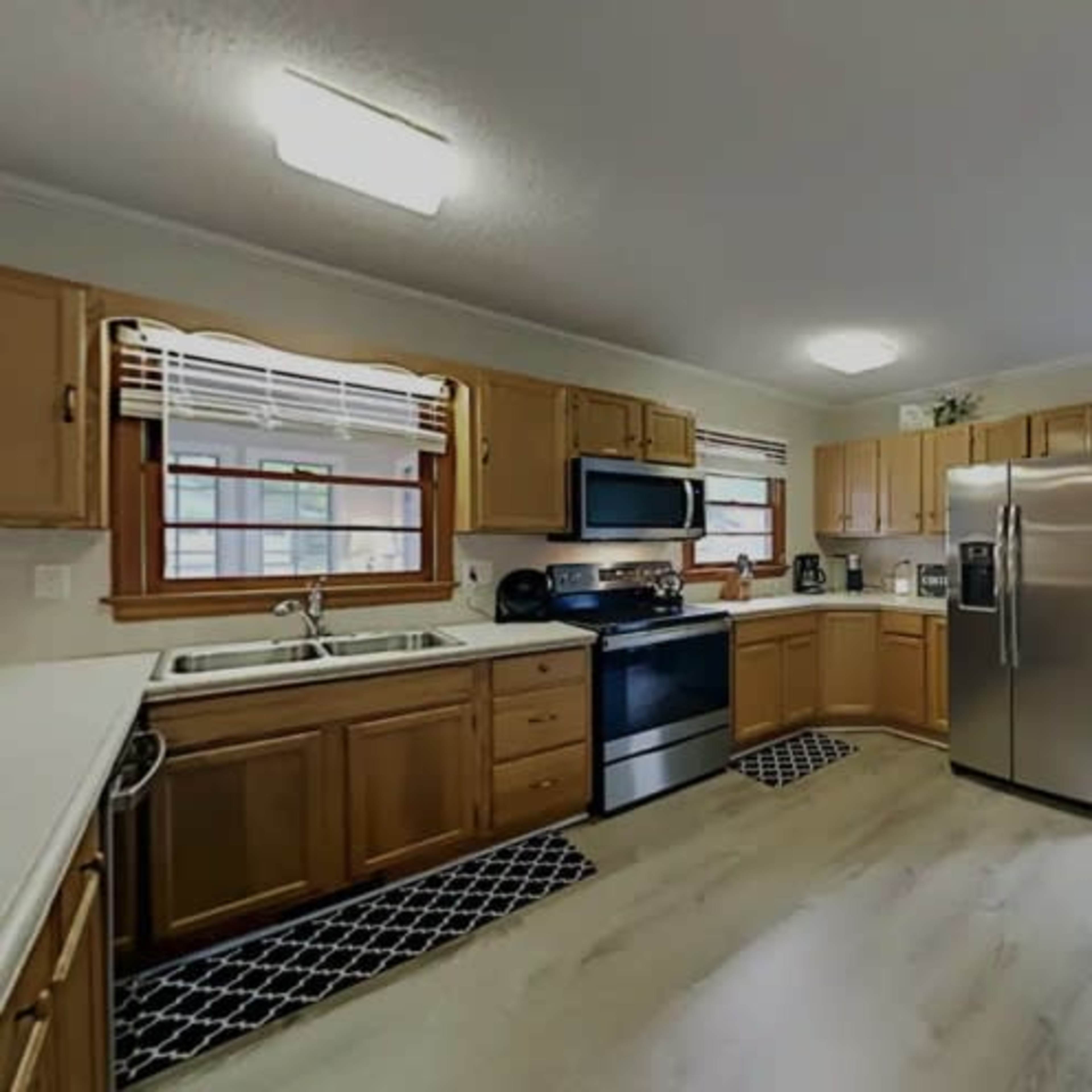 A kitchen features wooden cabinets, a stainless steel refrigerator, a black stove, and a white countertop with a window overlooking the outside.