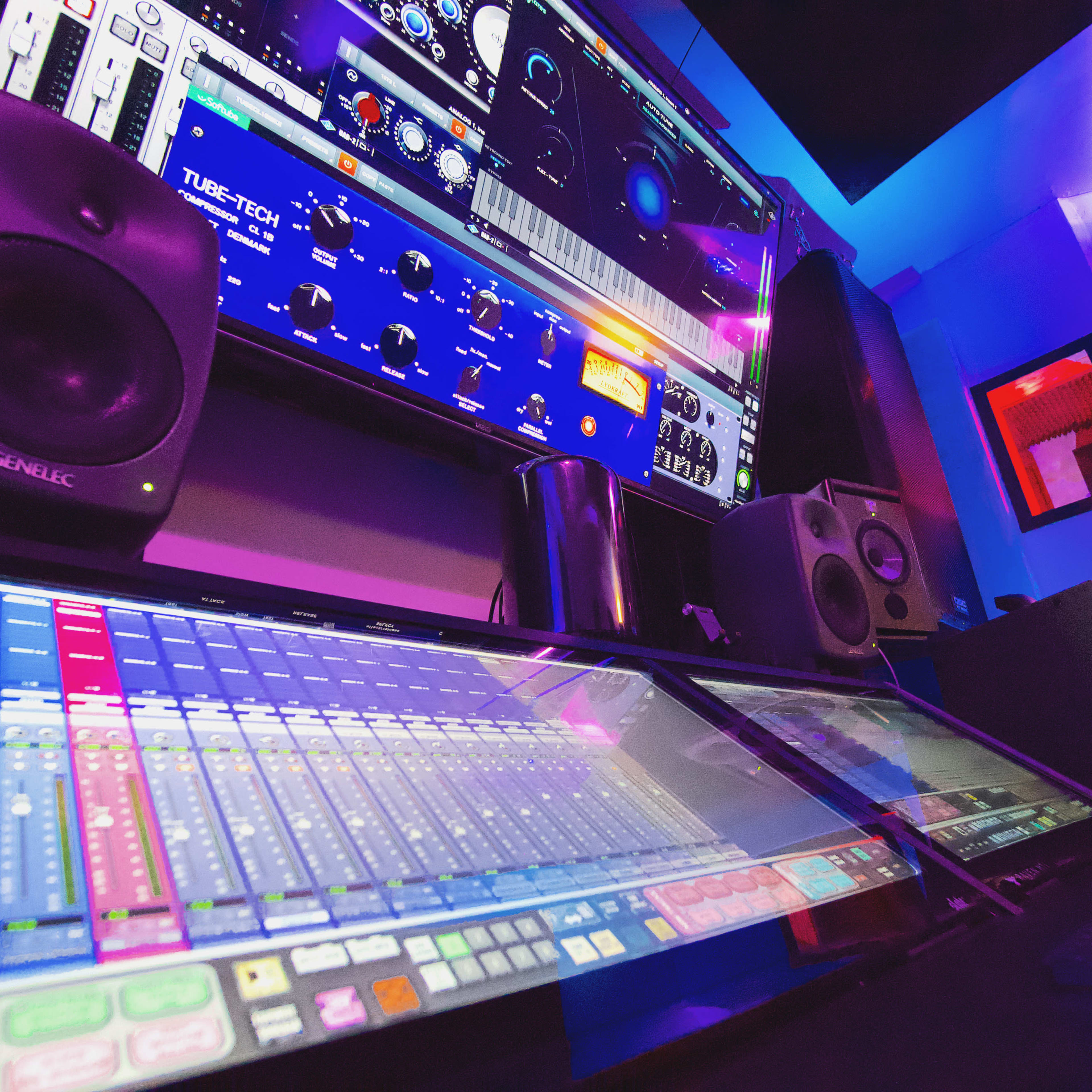 A music studio with computer monitors displaying audio mixing software, professional speakers, and various audio equipment along the walls.