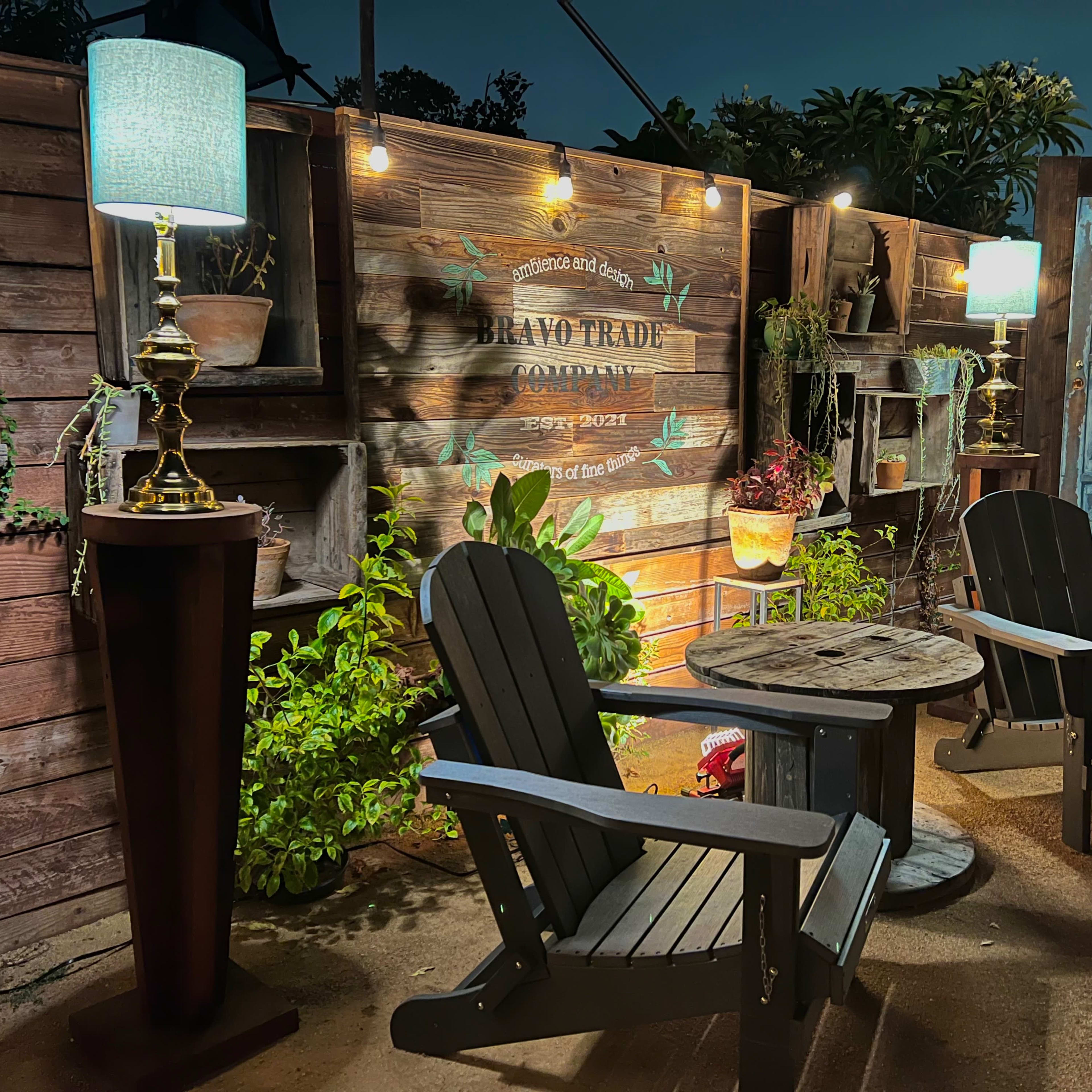 Two wooden Adirondack chairs sit beside a small table under illuminated lamps in a garden setting with a rustic wooden wall and greenery.