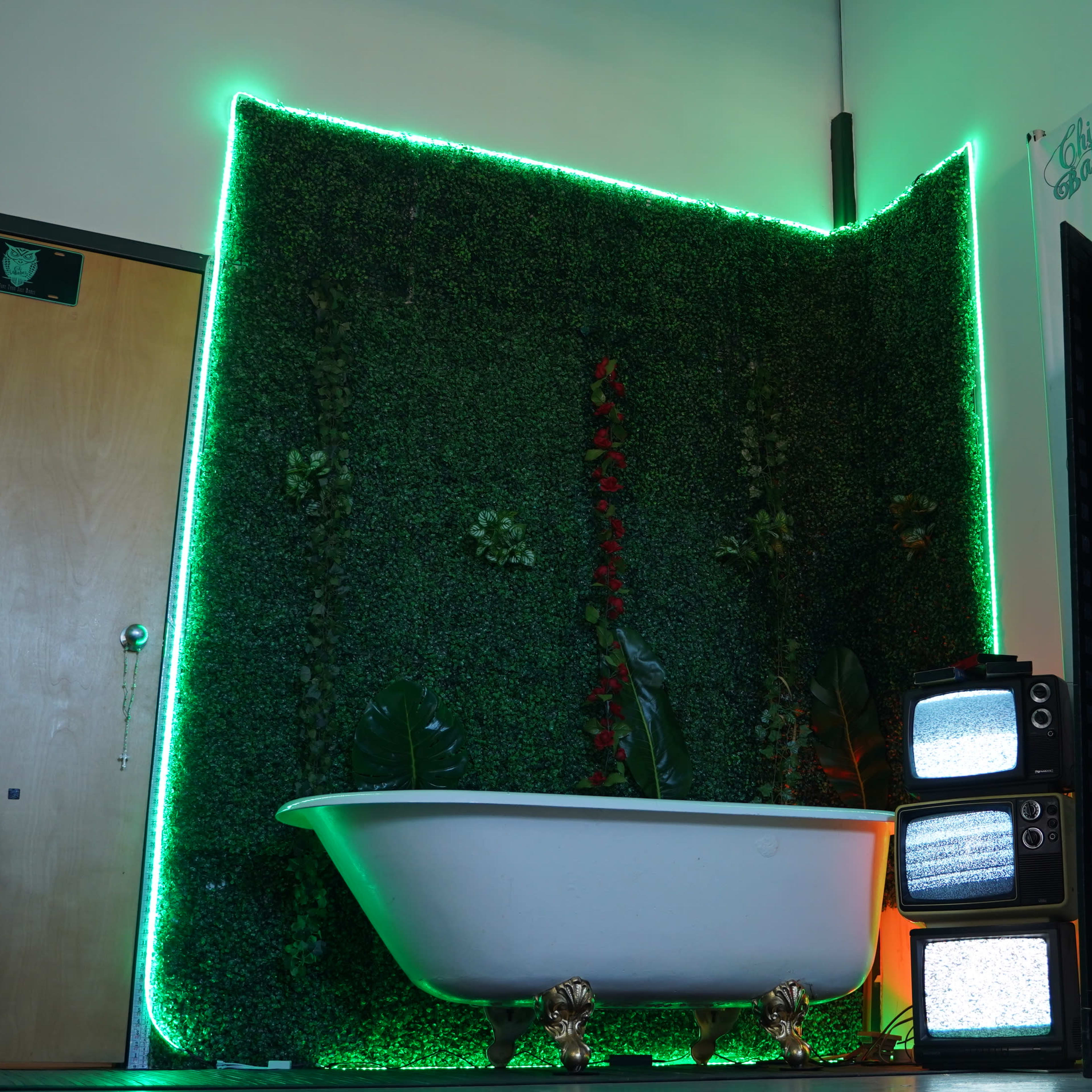 A vintage clawfoot bathtub sits in front of a green wall adorned with plants and illuminated by neon lights, next to a stack of retro television sets.