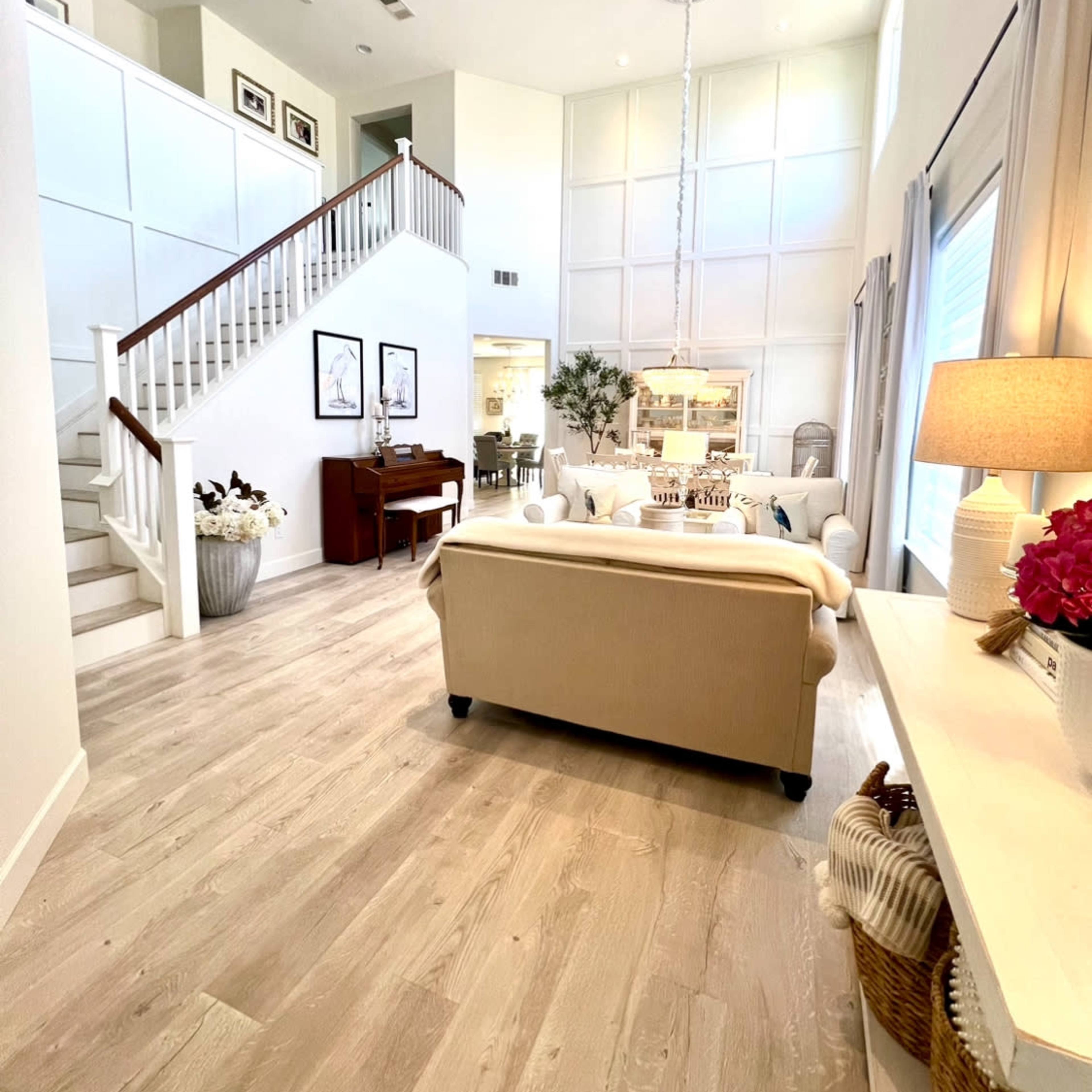 A spacious living room features a light-colored sofa, a piano, and a staircase leading to an upper level, with large windows allowing natural light to fill the space.