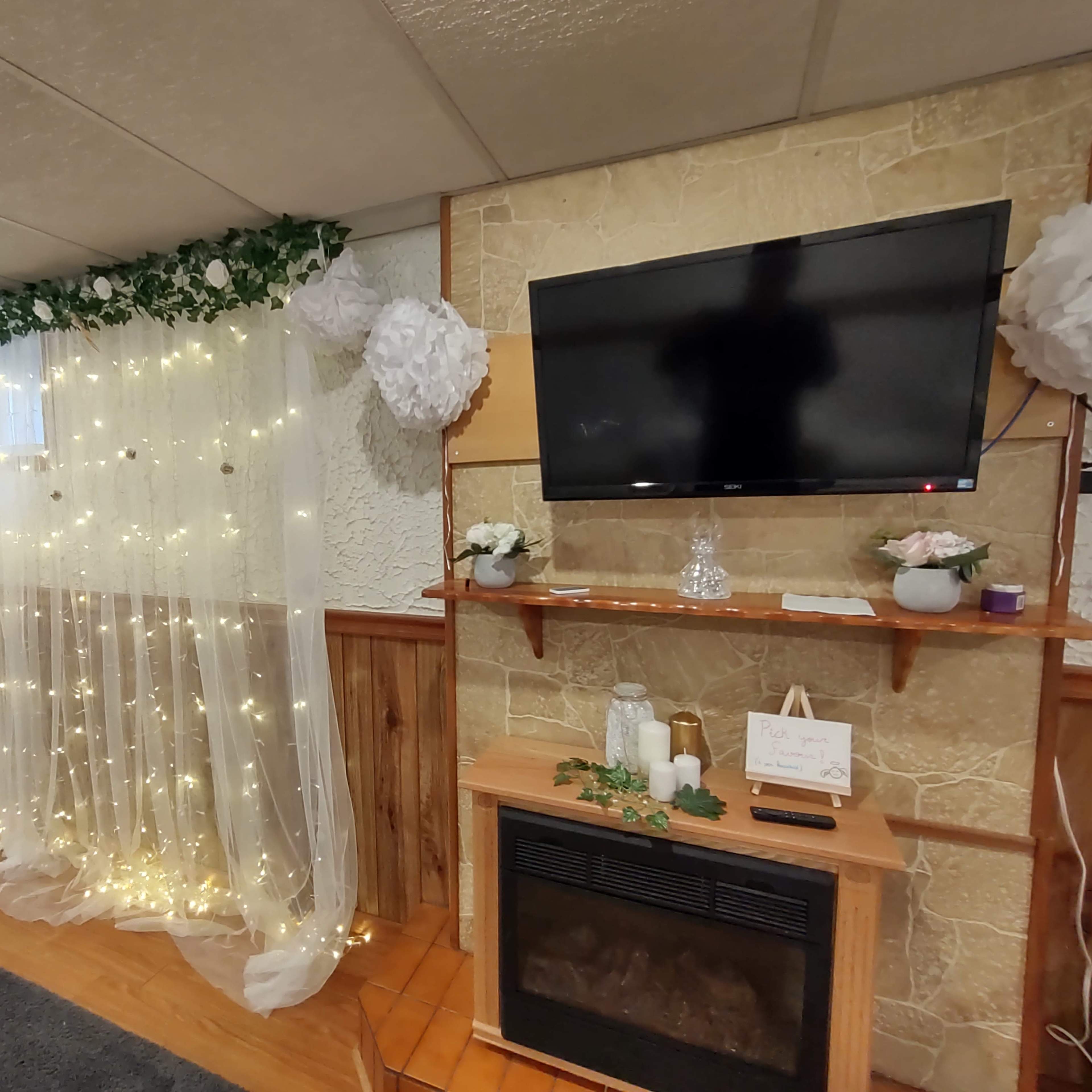 A basement room with a wall draped in sheer curtains adorned with fairy lights, a TV mounted on a stone wall, and a wooden shelf with decorative items above a fireplace.