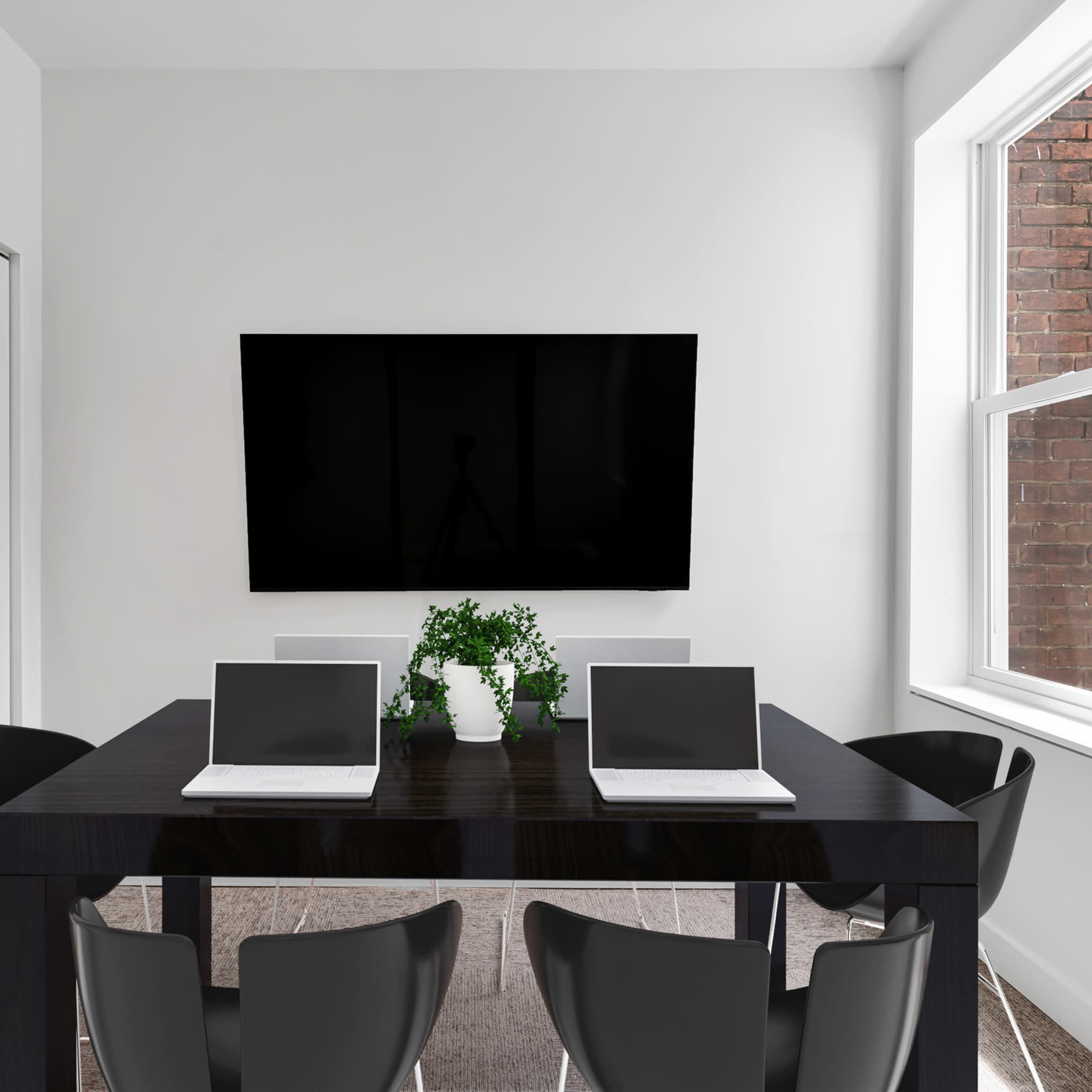 A modern meeting room features a black table with four chairs, two laptops, a green plant centerpiece, and a large television mounted on the white wall.