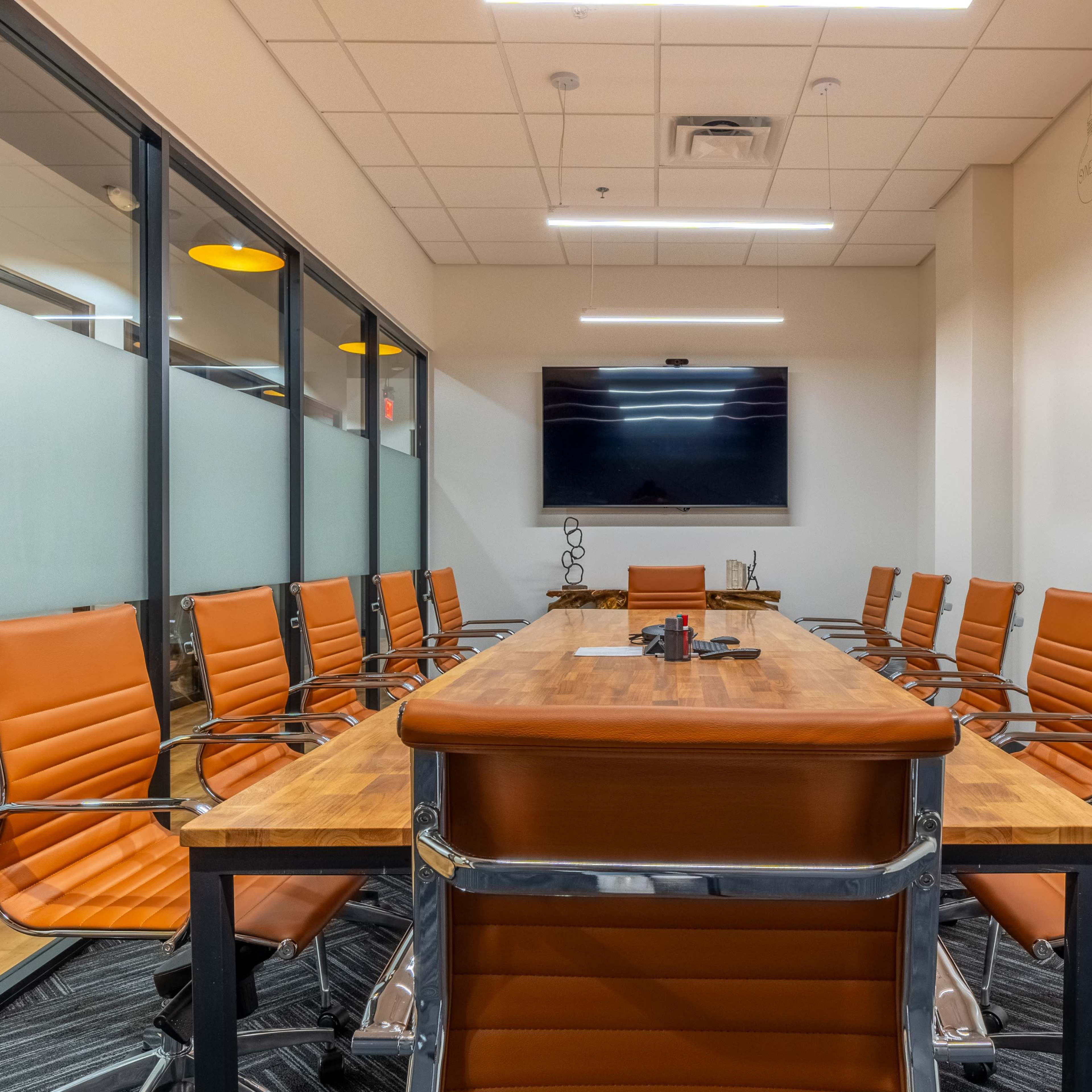 A modern conference room features a long wooden table surrounded by orange chairs and a wall-mounted television.