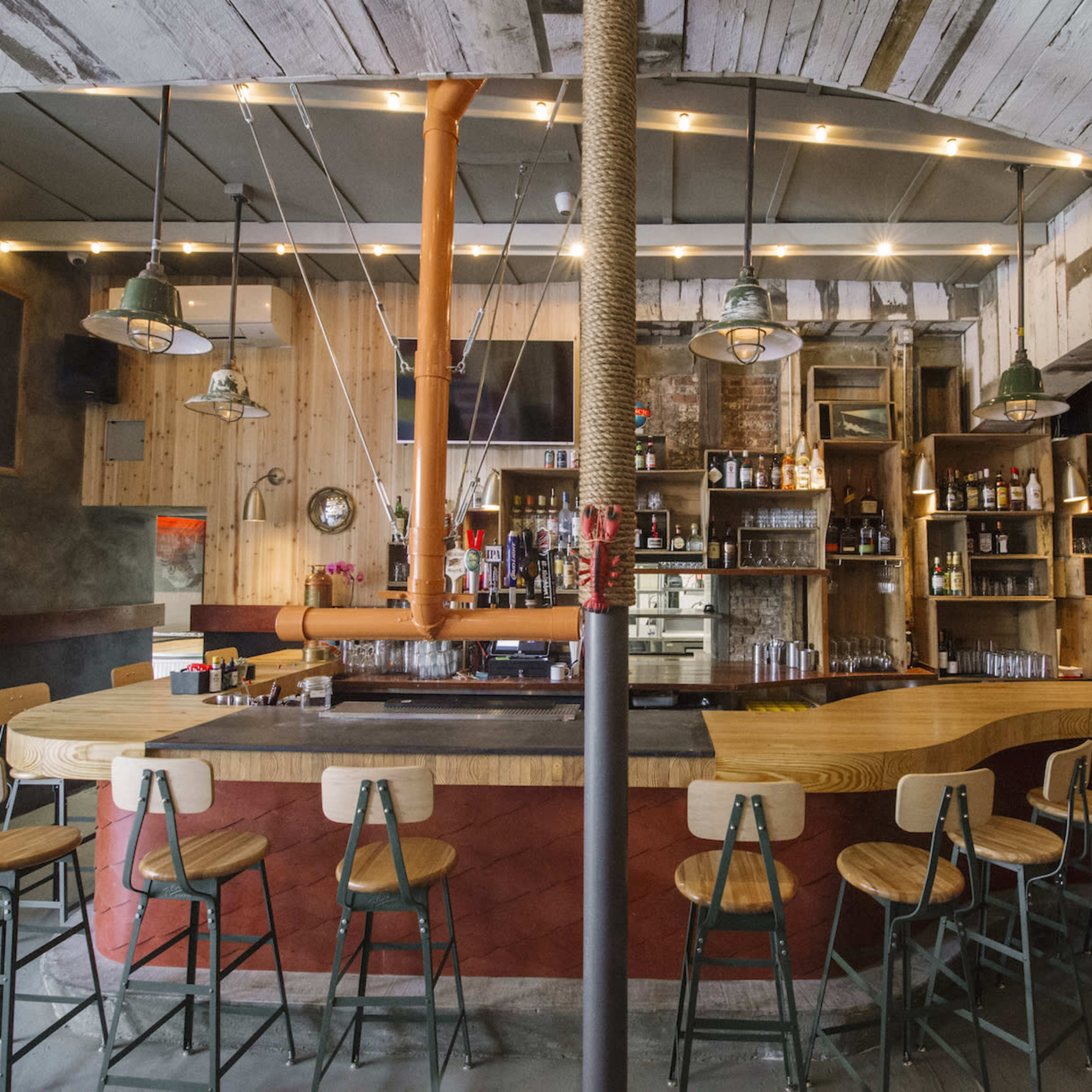 A rustic bar with wooden stools arranged around a curved counter, illuminated by pendant lights amidst a backdrop of shelves stocked with various bottles.