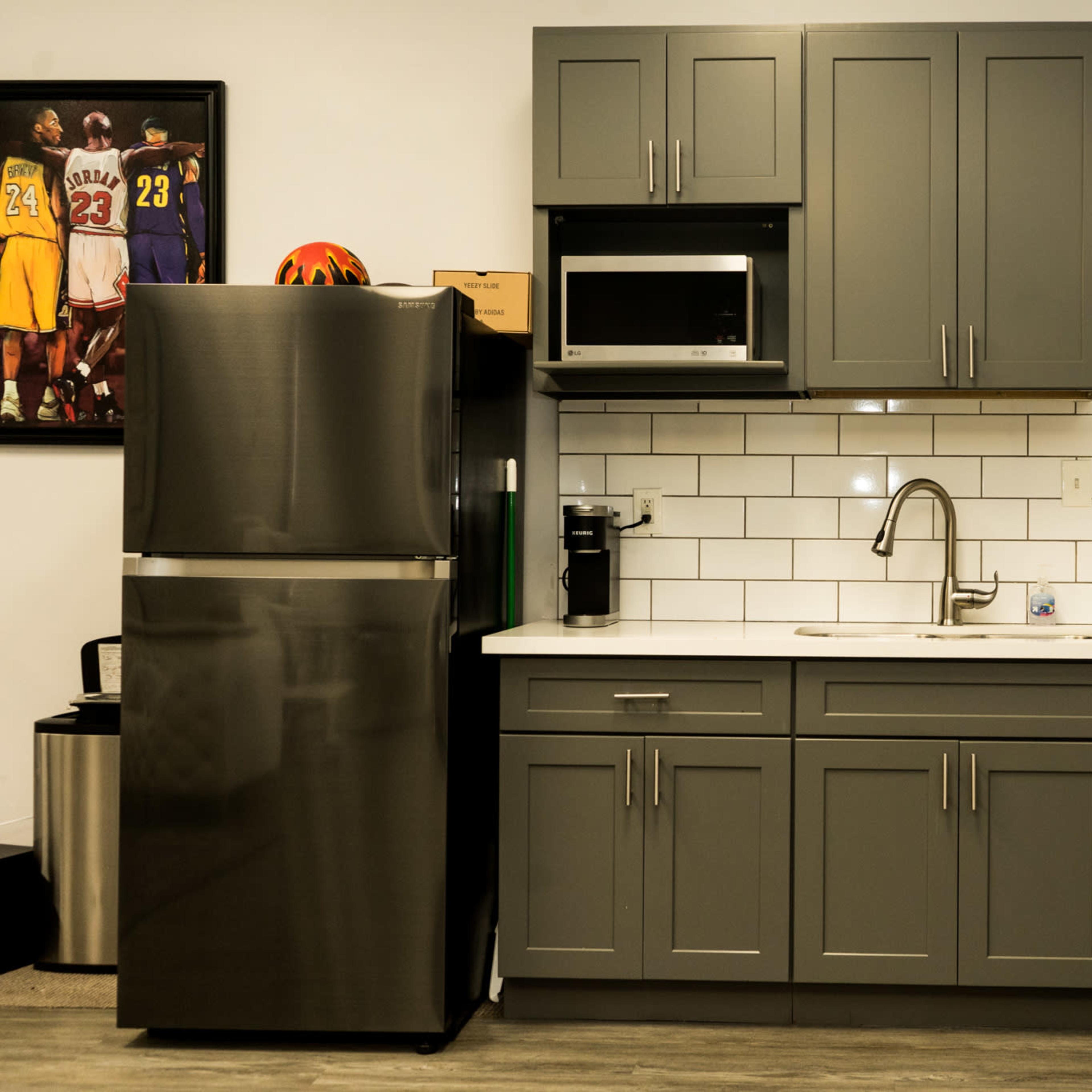 A modern kitchen area featuring gray cabinetry, a stainless steel refrigerator, a microwave, and a white tile backsplash.