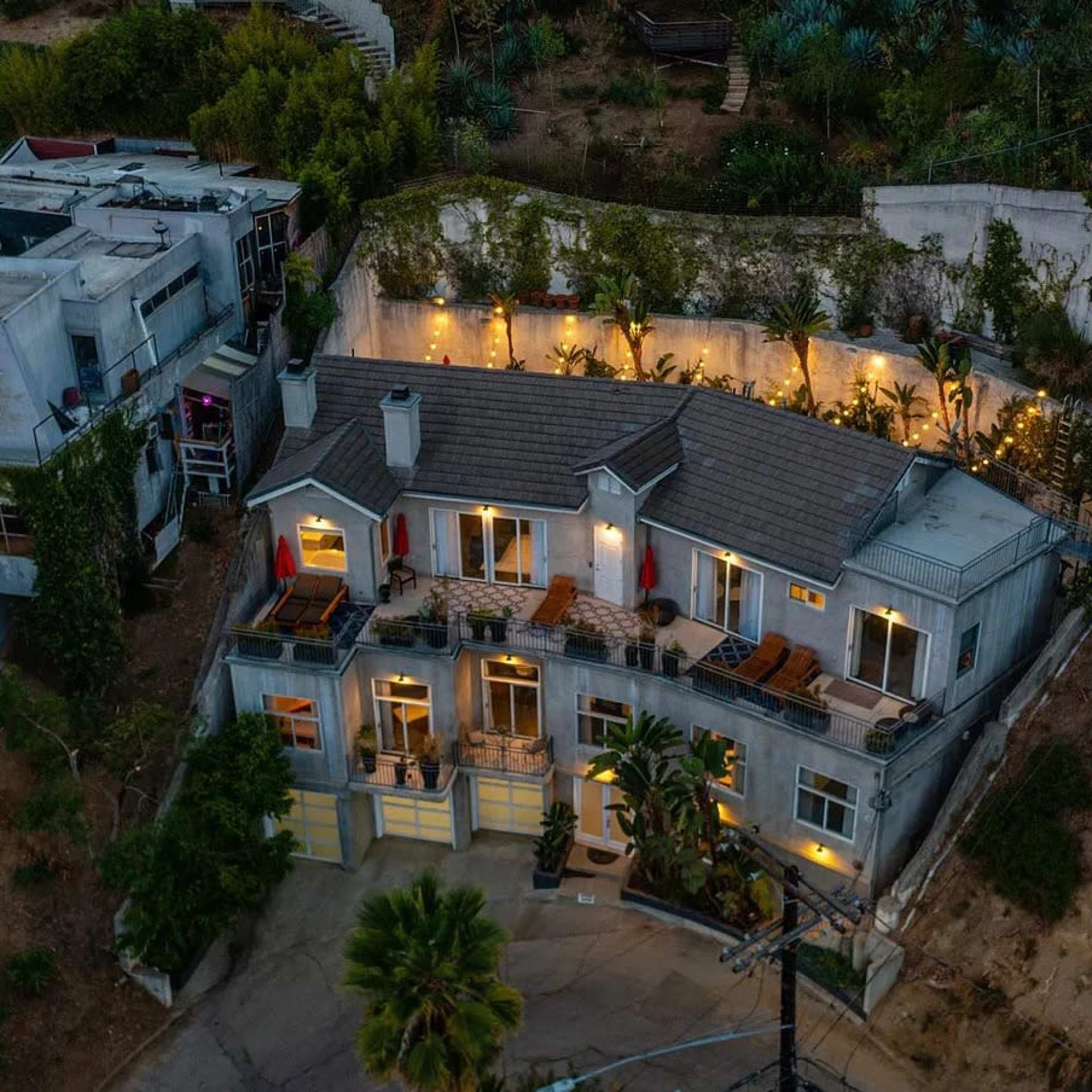 A modern house situated on a hillside, surrounded by greenery and featuring outdoor lighting on its terraces.