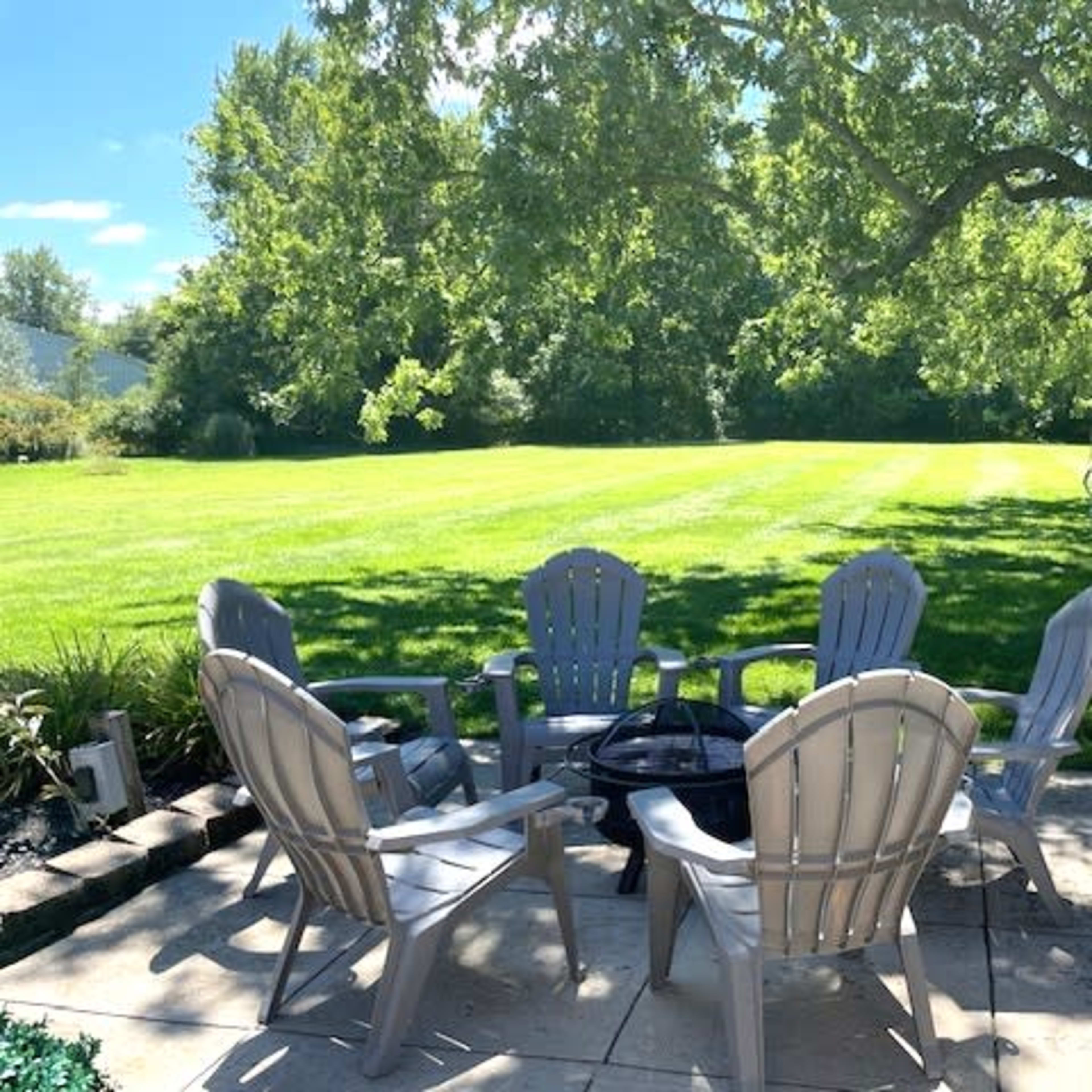 A patio area features six gray chairs arranged around a fire pit, overlooking a large grassy field and shaded by trees.