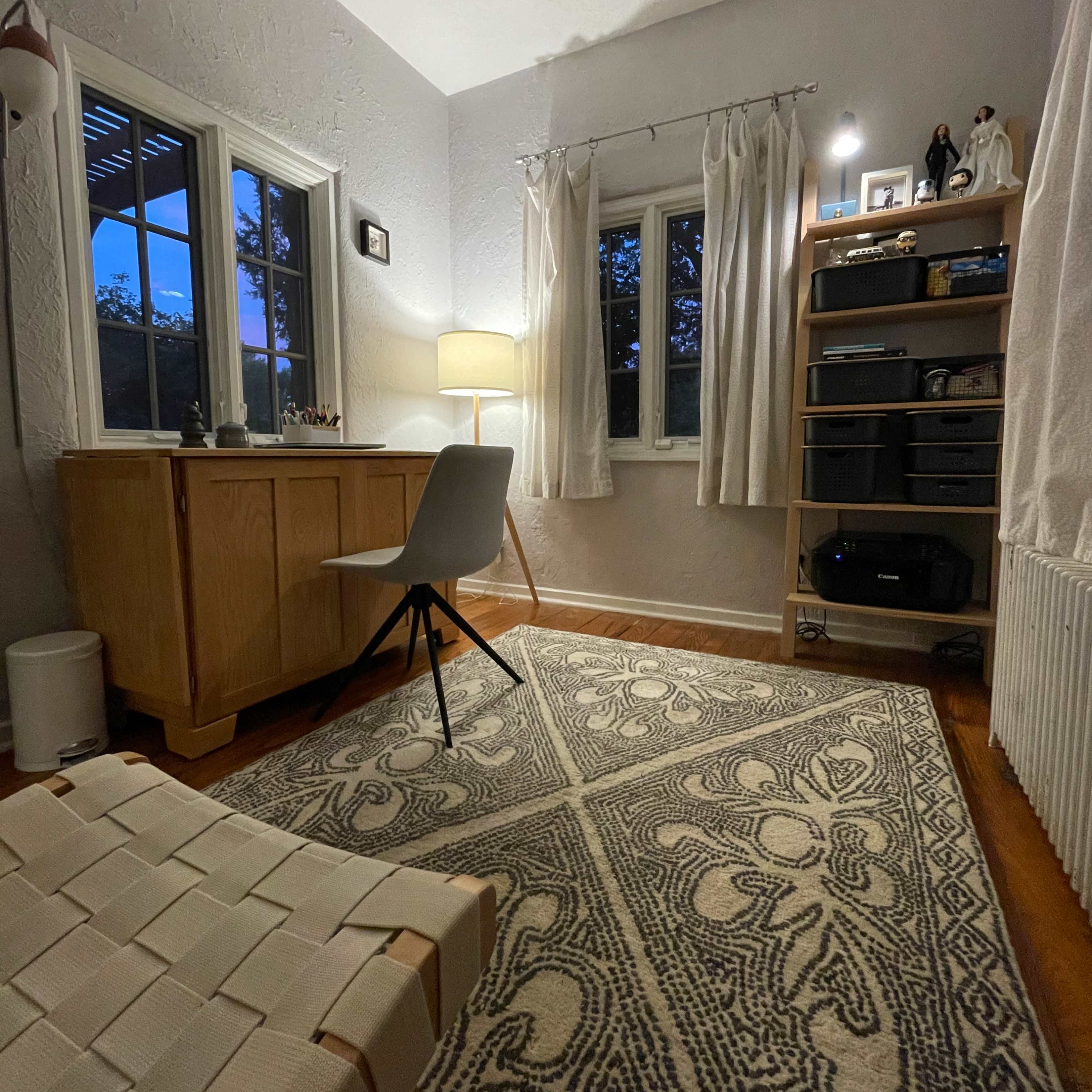 A well-lit room features a wooden desk with a chair, a bookshelf, and a patterned rug.