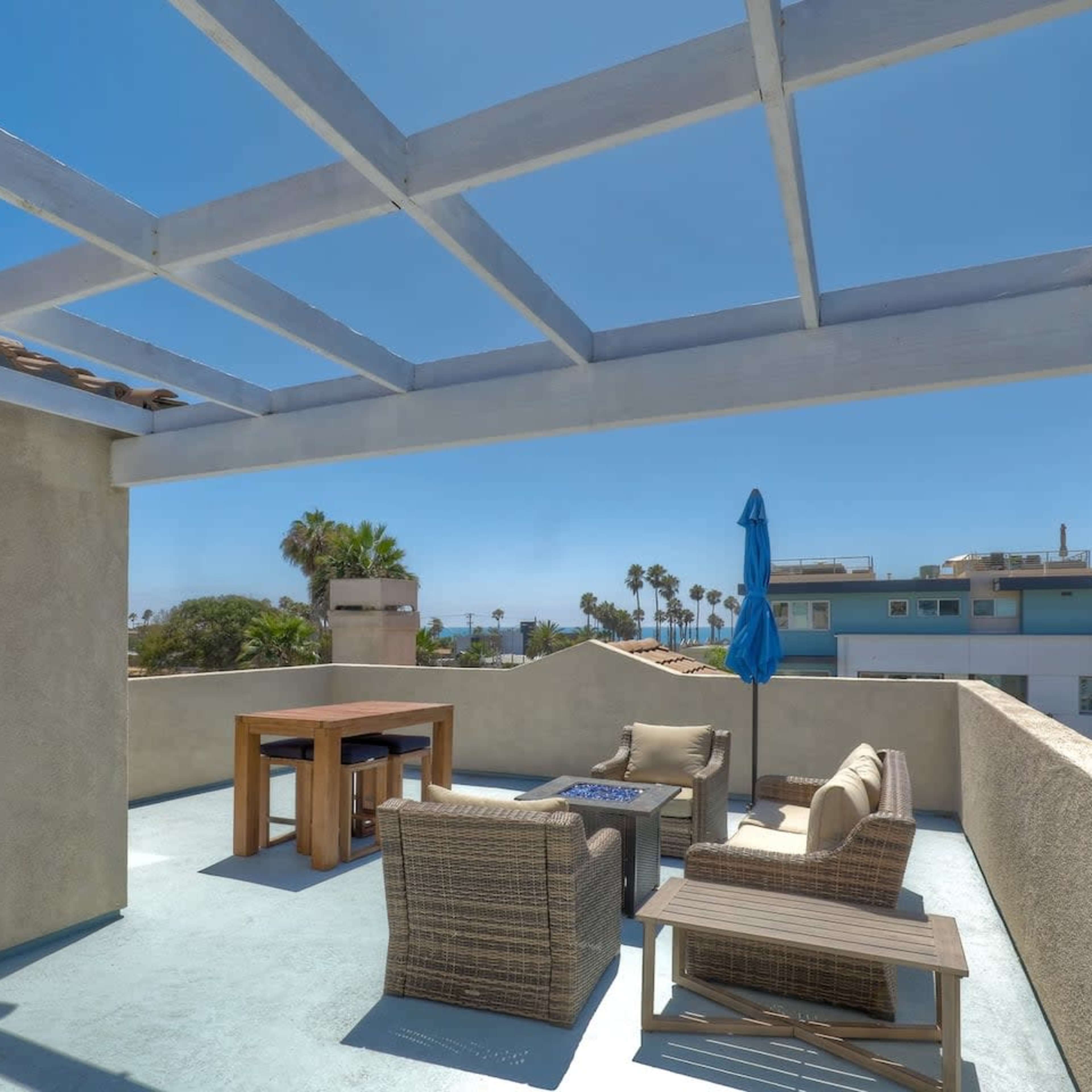 A rooftop terrace features a table and seating area beneath a pergola, with a blue umbrella and palm trees visible in the background.