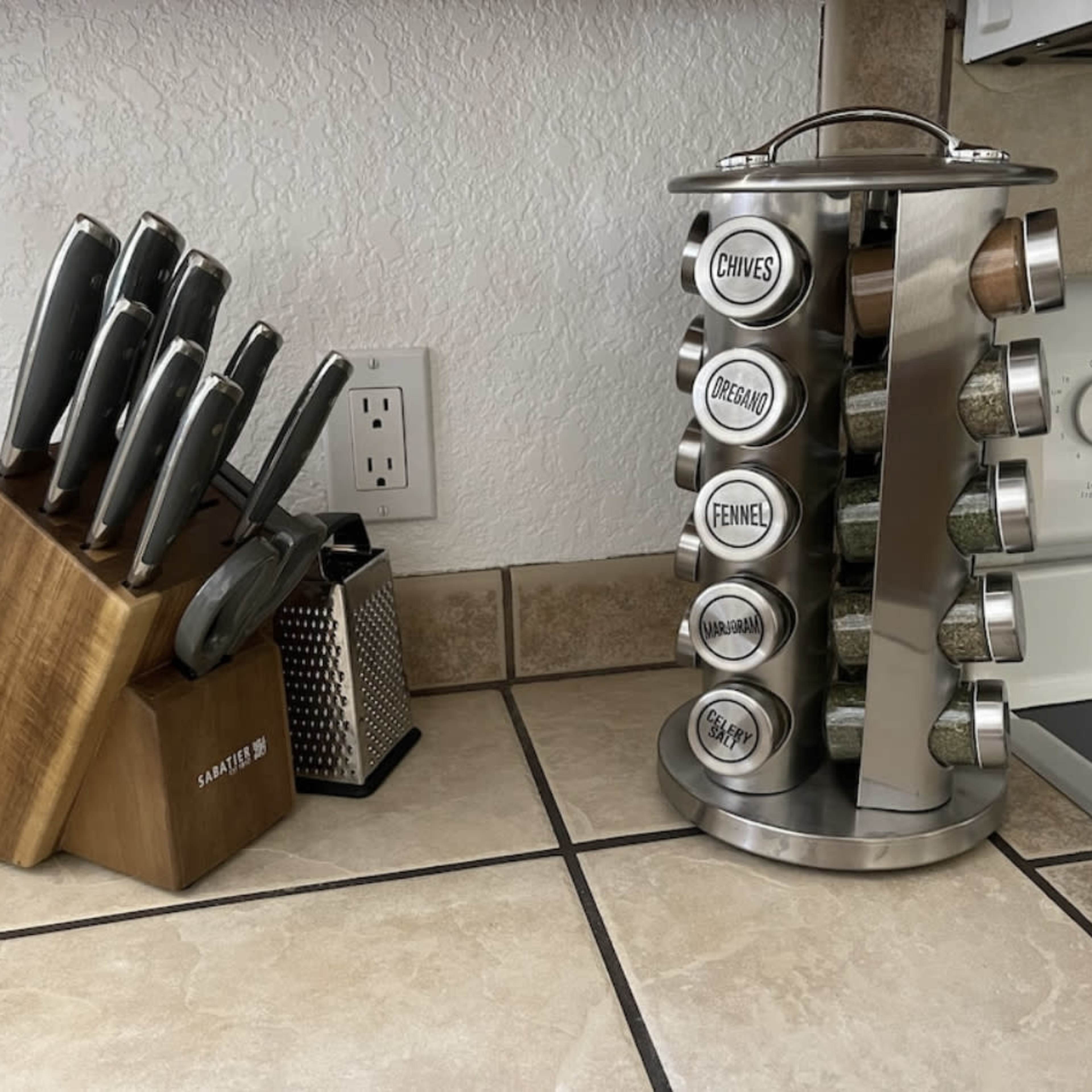 A wooden knife block with several knives on the left and a stainless steel spice rack holding labeled spice jars on the right, situated on a tiled kitchen counter.