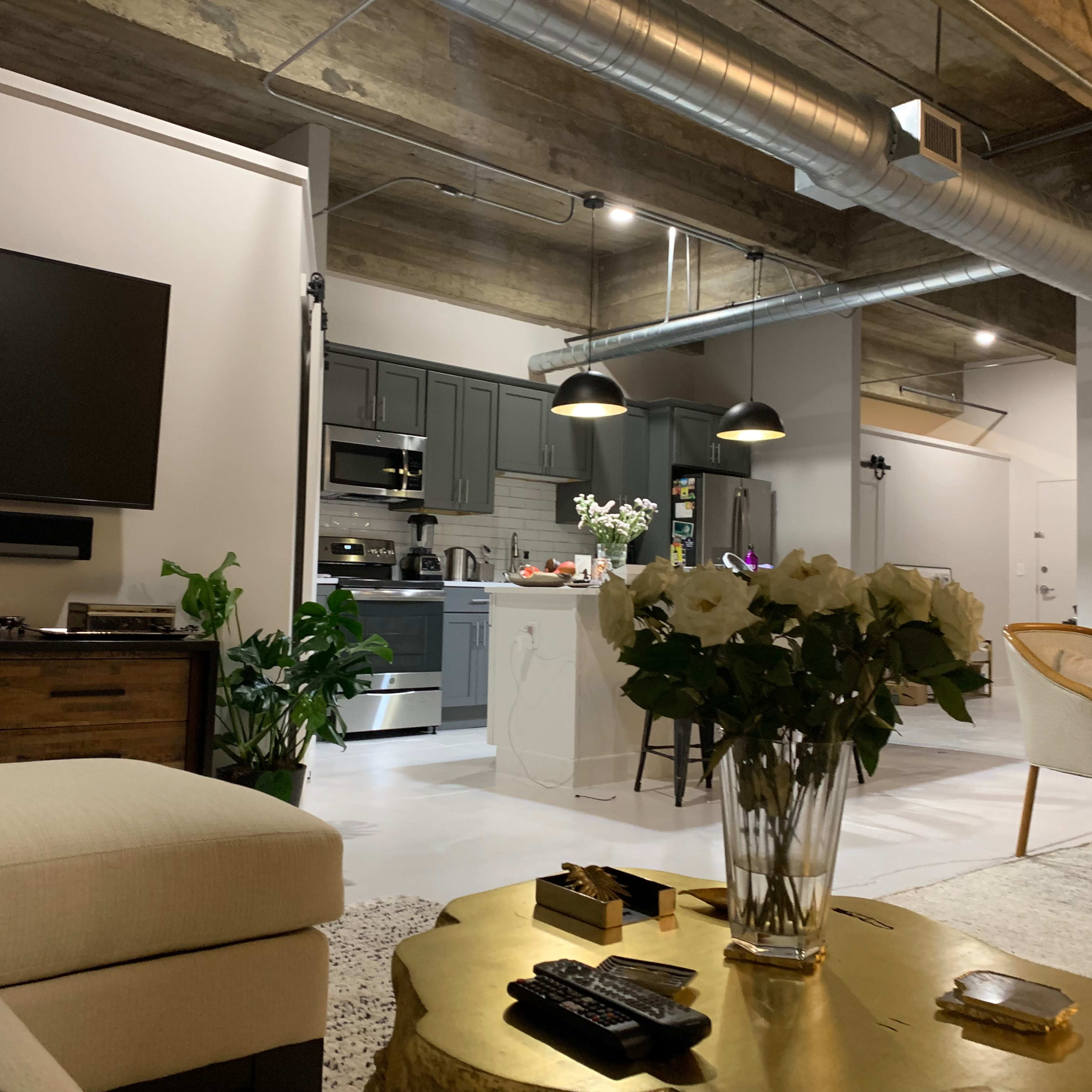 A modern, open-concept living space featuring a comfortable couch, a television mounted on the wall, a kitchen in the background, and a dining area with a decorative table.