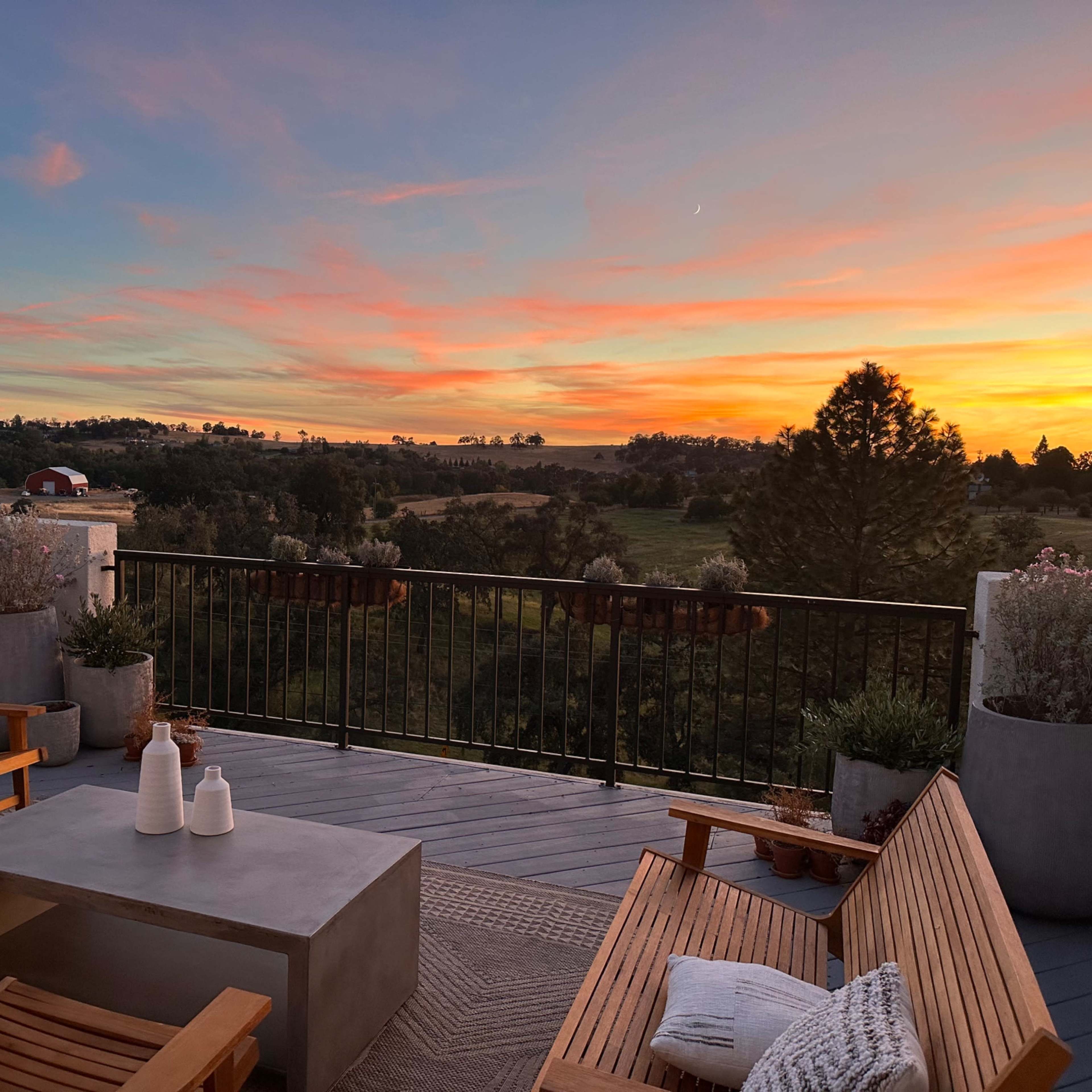 A wooden deck is furnished with chairs and a table, overlooking fields and a colorful sunset in the distance.