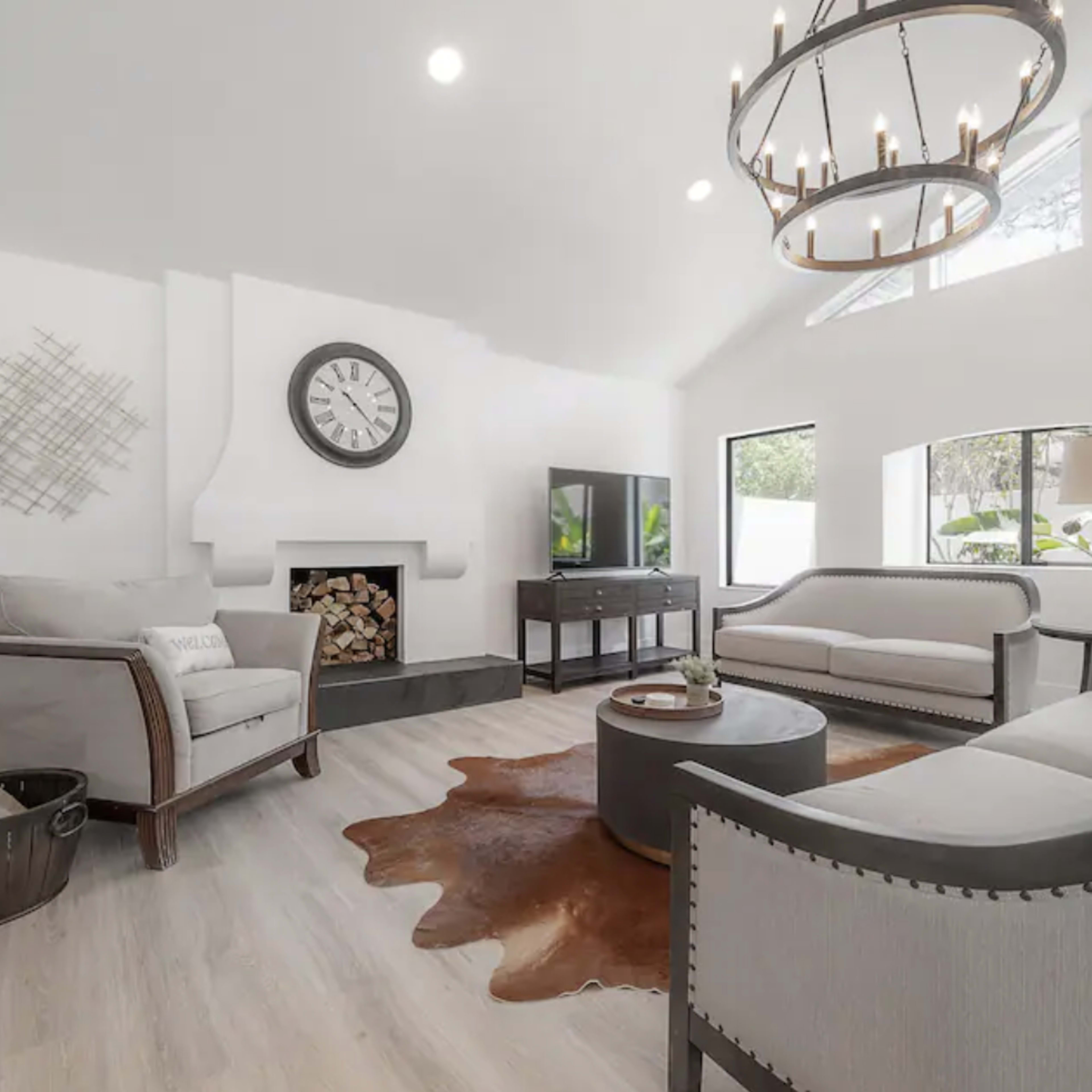 The living room features light-colored walls, a modern chandelier, a fireplace with stacked logs, and minimalist furnishings, including a circular coffee table and two sofas.