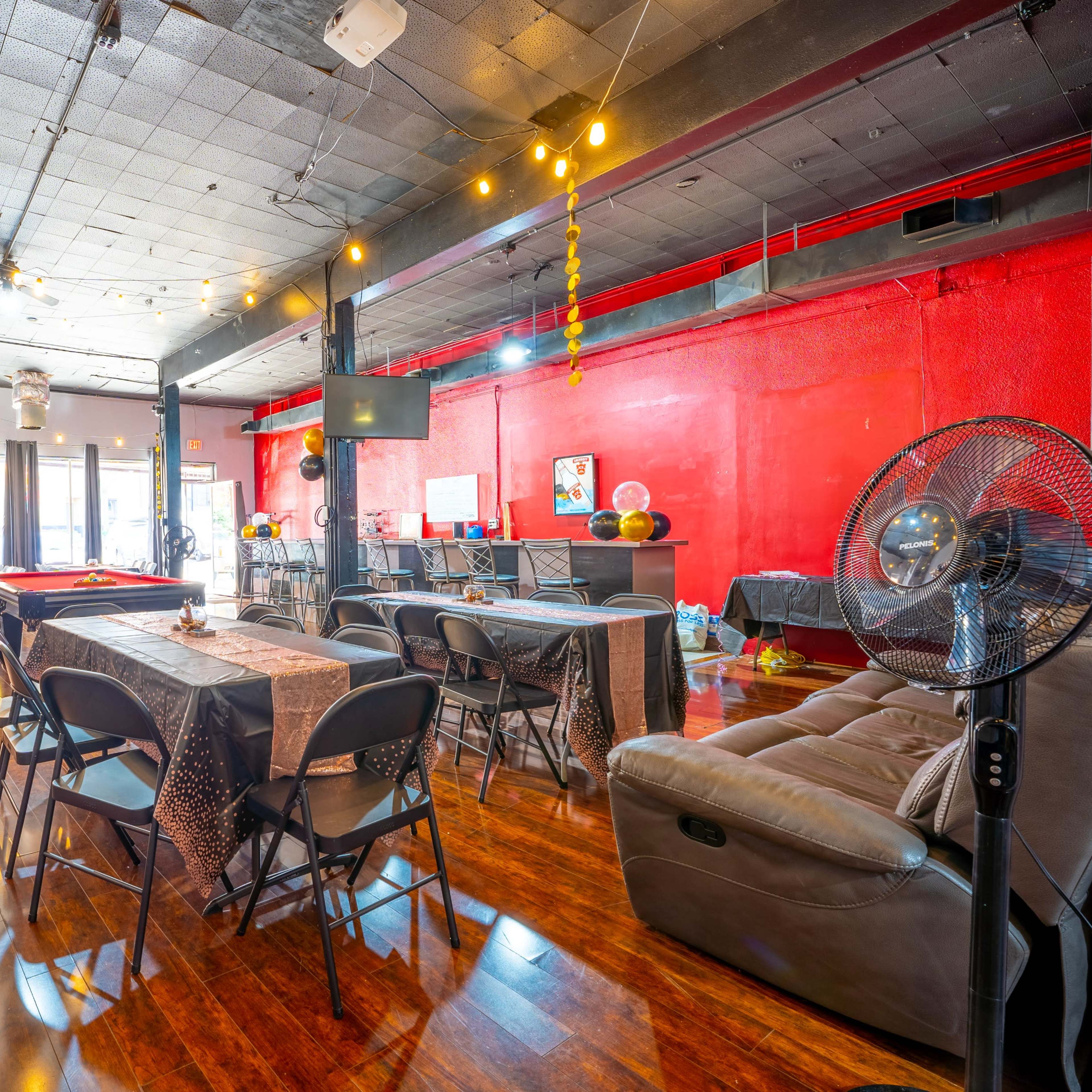A spacious event venue with tables and chairs arranged, colorful decorations, and a bar area in the background.